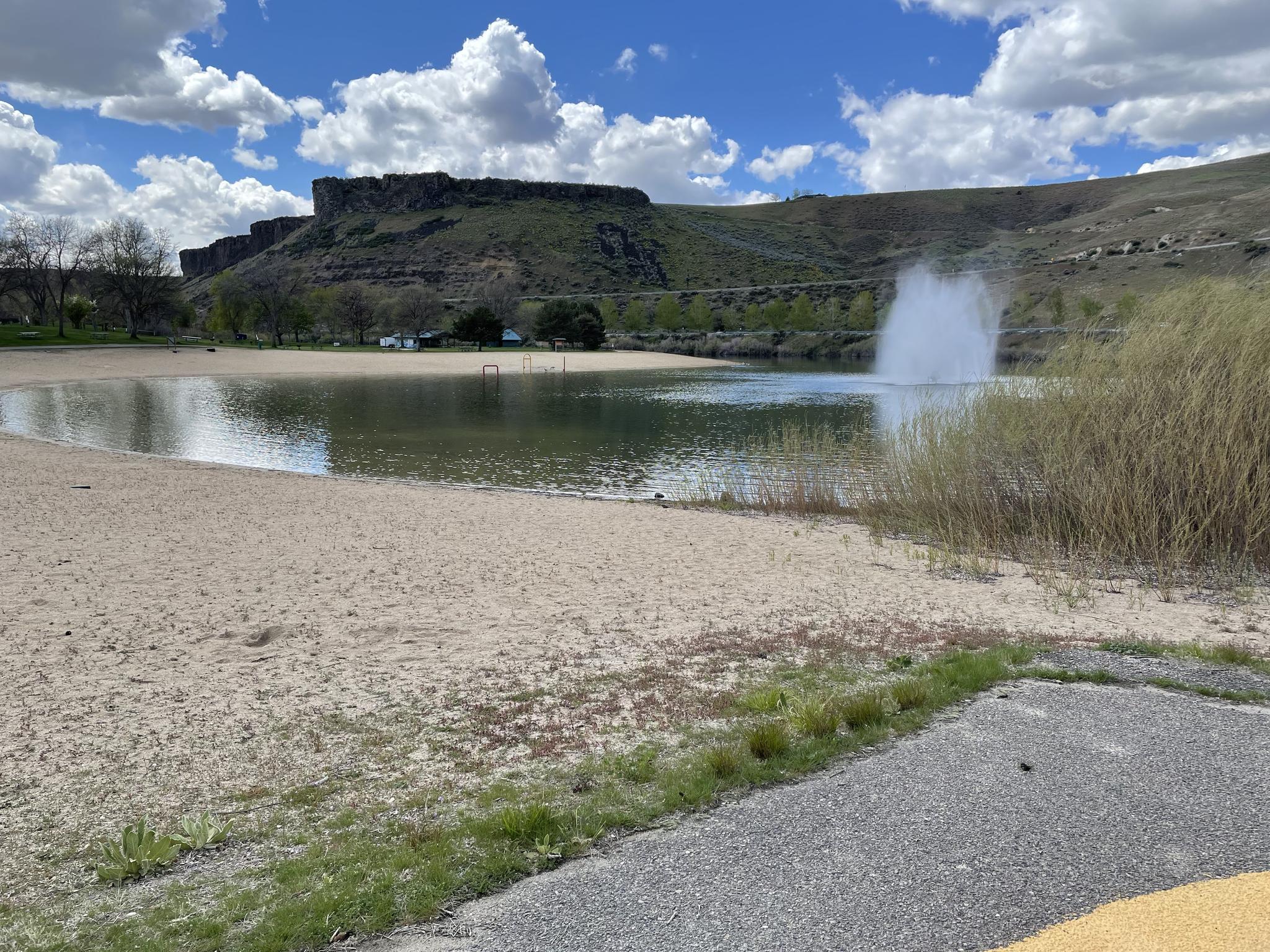 A sandy beach and lake with a fountain, backed by green hills under a cloudy sky.