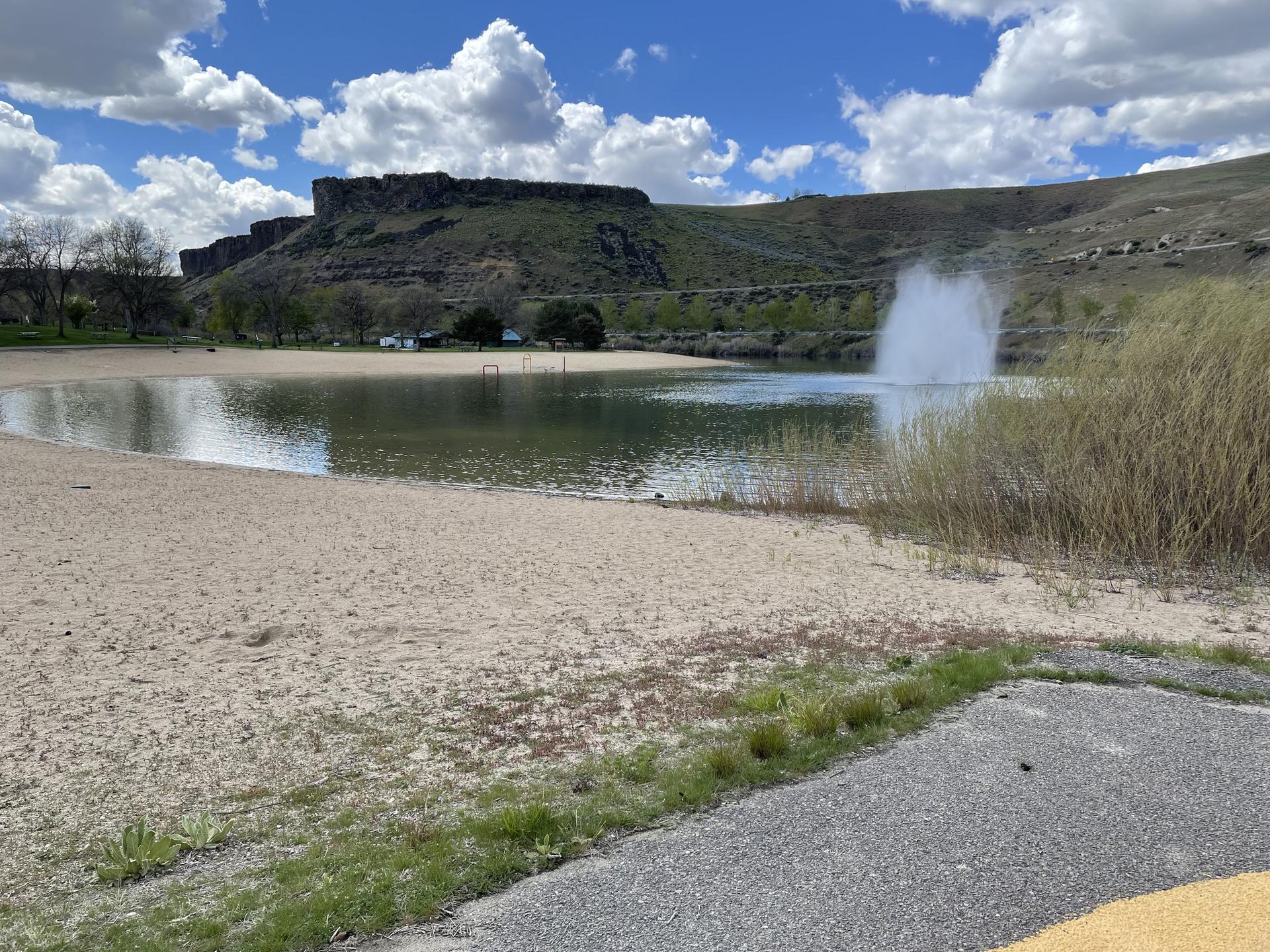A sandy beach and lake with a fountain, backed by green hills under a cloudy sky.