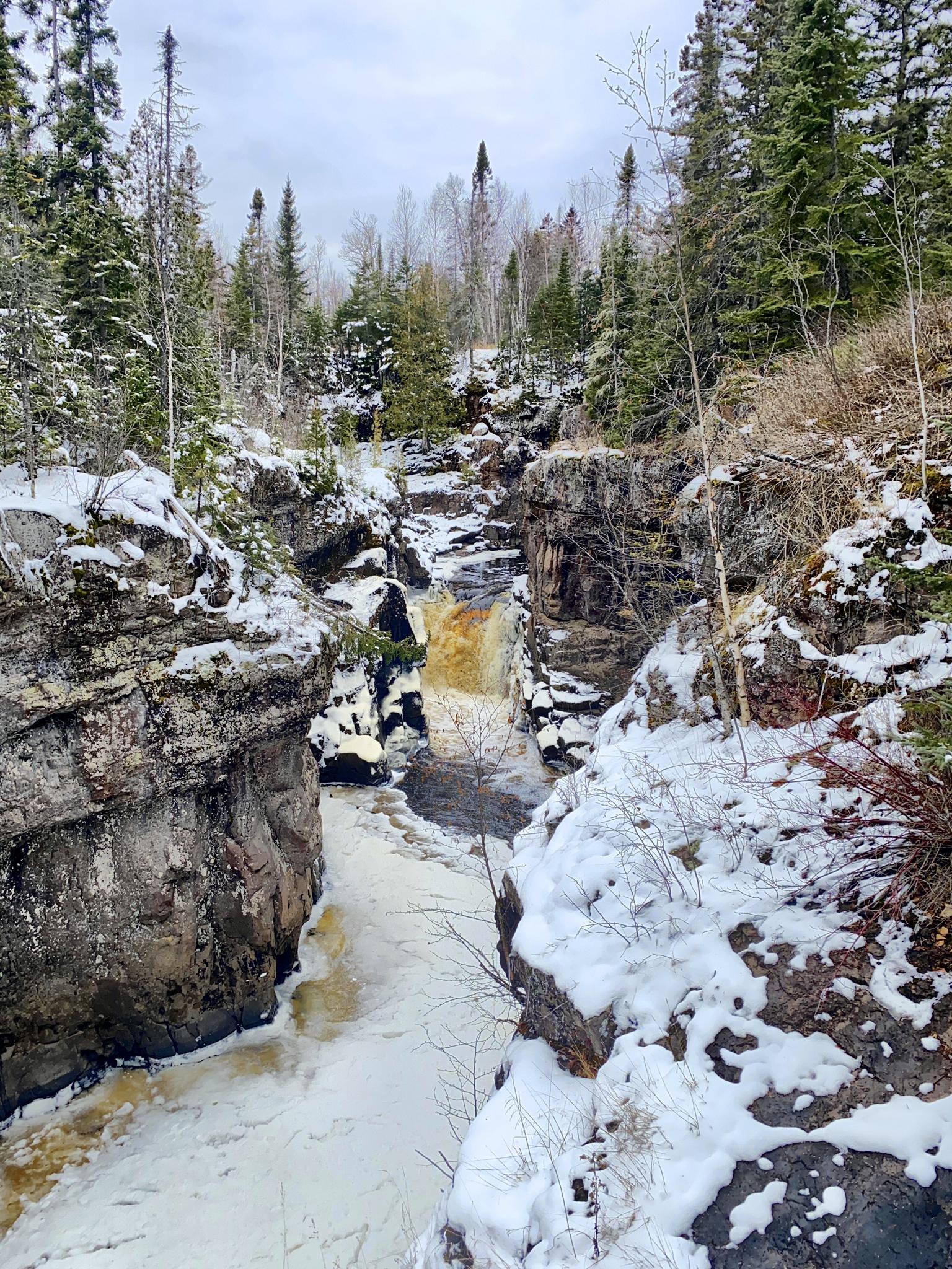 A brown waterfall plunges into a snowy, rocky gorge surrounded by evergreen trees.