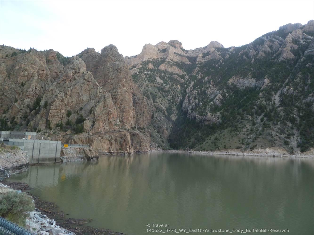 Reservoir and concrete dam at the base of rugged mountains under a cloudy sky.