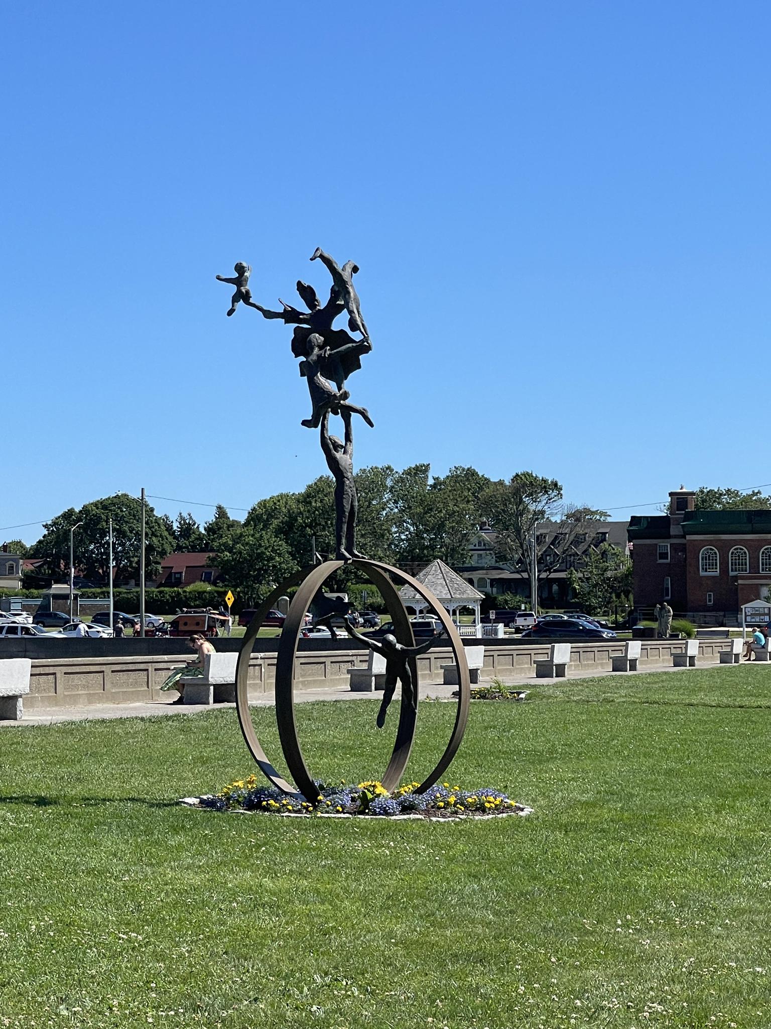 Bronze sculpture of balancing figures on rings in a sunny green park.