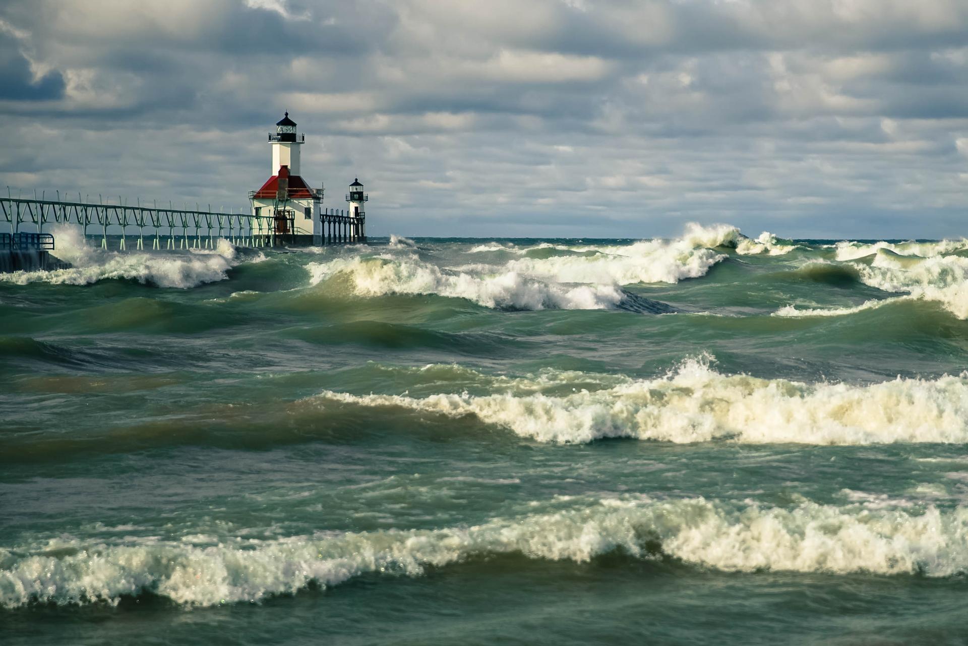 A lighthouse on a pier with large, crashing waves under a cloudy sky.
