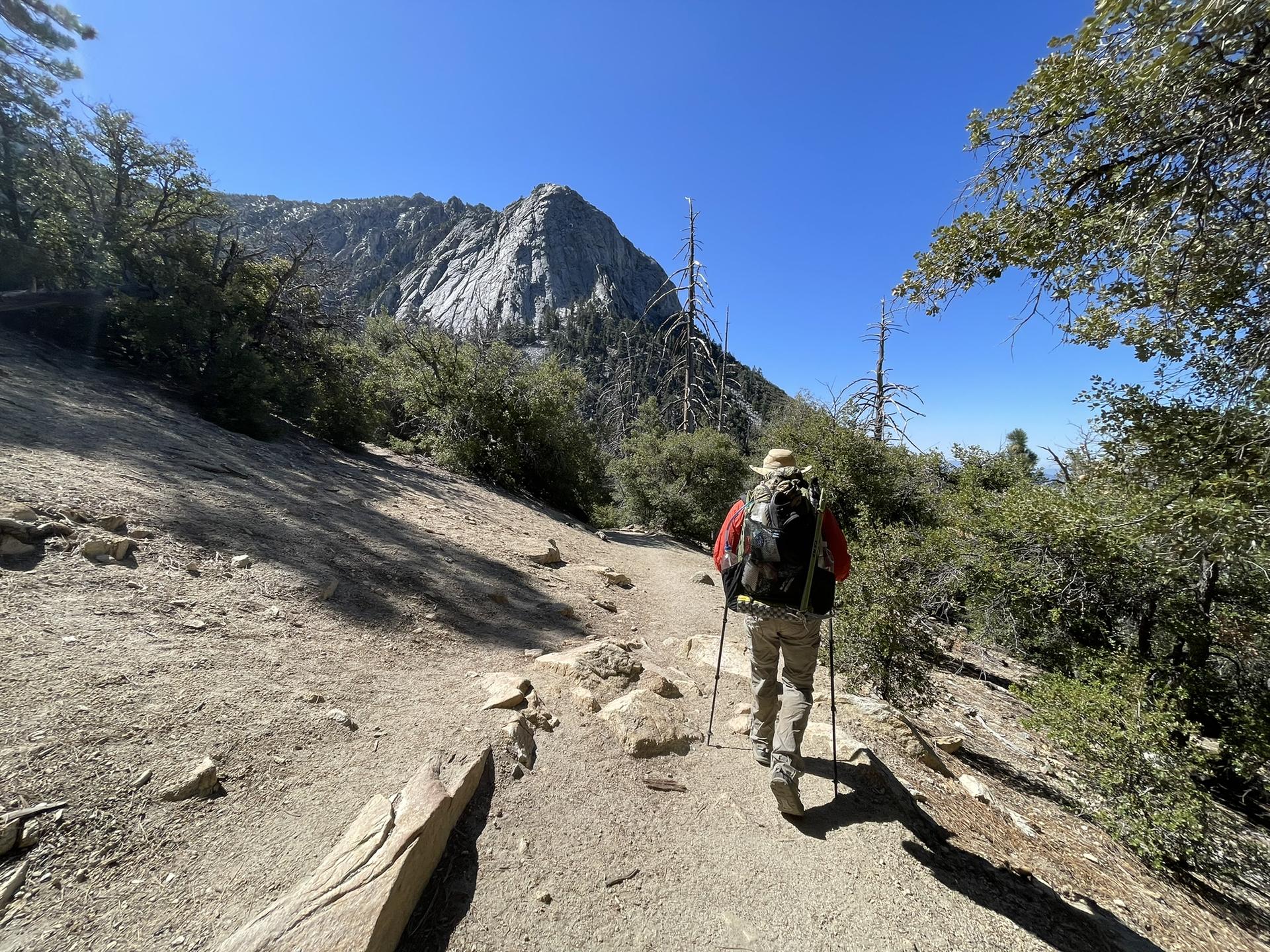 A hiker with a backpack and poles walks on a dirt trail towards a peak.