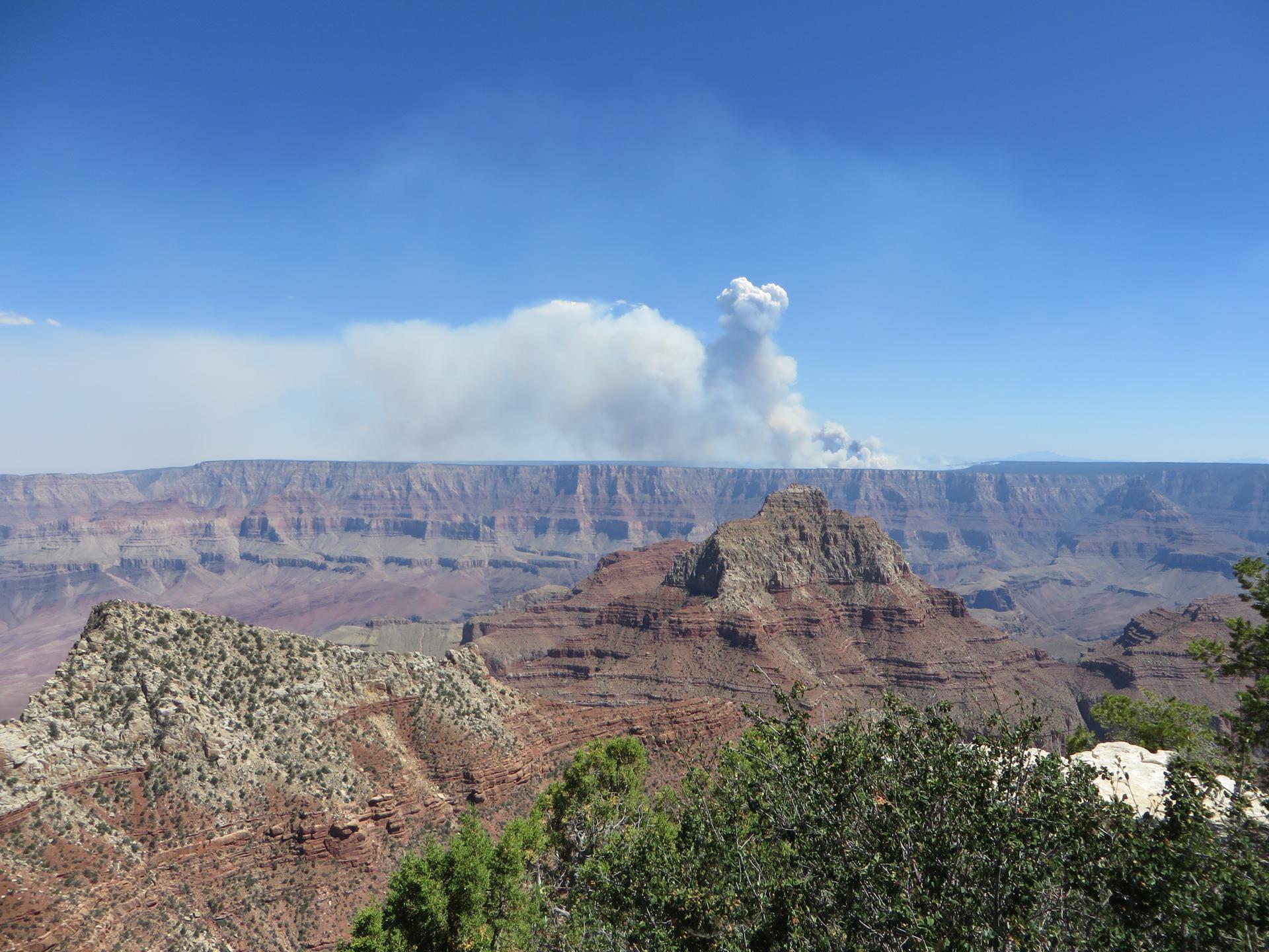 Vast canyon landscape under a blue sky with a large smoke plume.