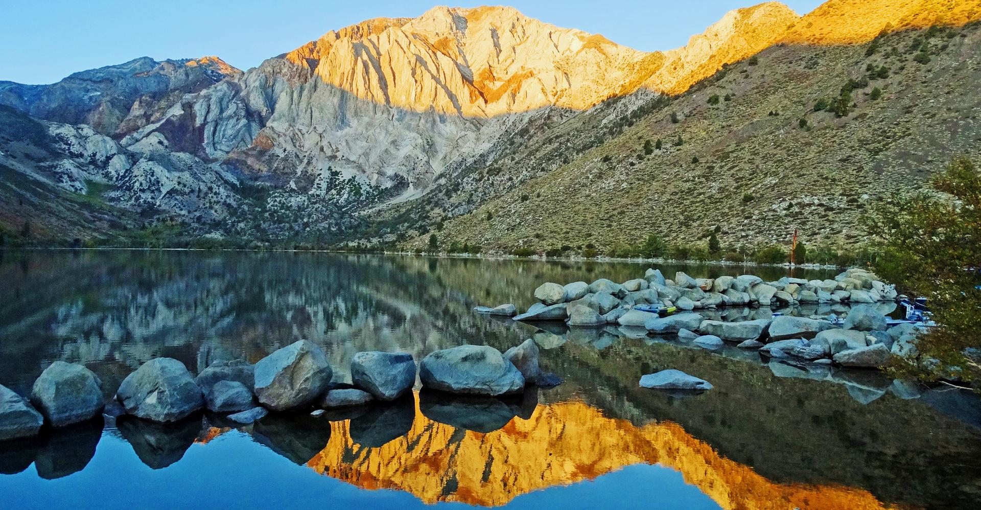 Golden light illuminates rugged mountains reflecting in a calm lake with rocks.