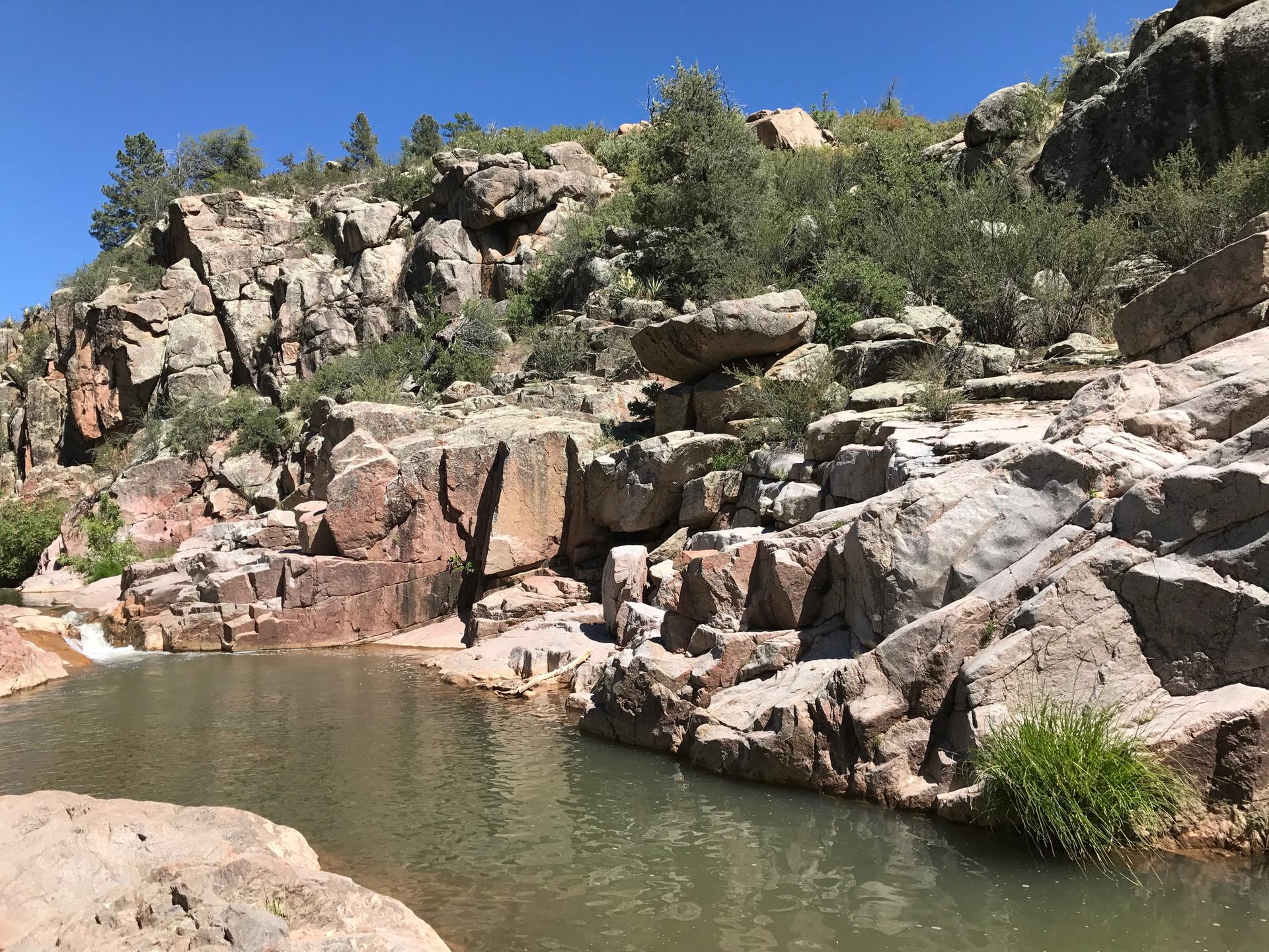 Sunny view of a natural pool amidst rocky cliffs and green bushes.