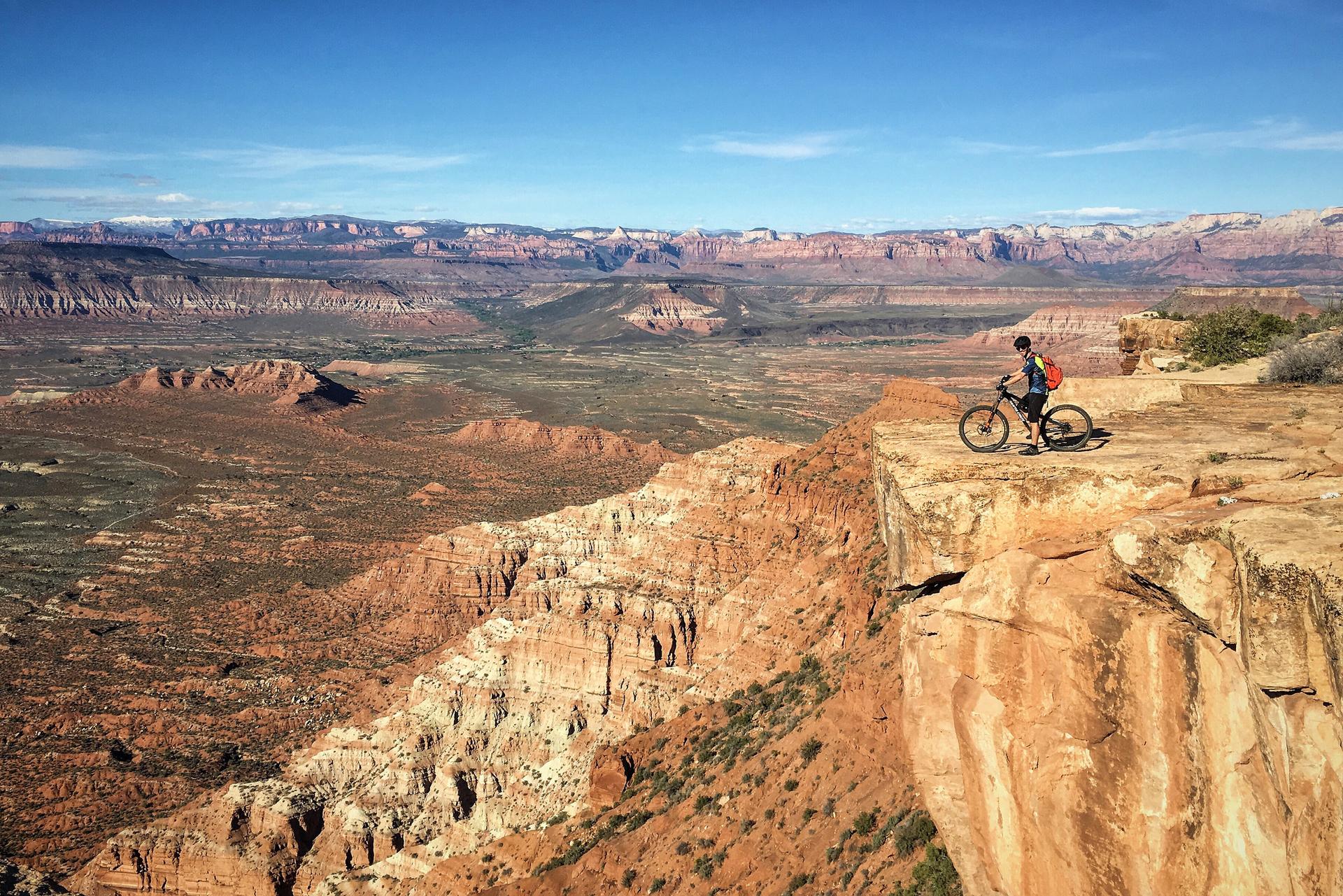 Mountain biker stands on a cliff edge overlooking a vast desert landscape.