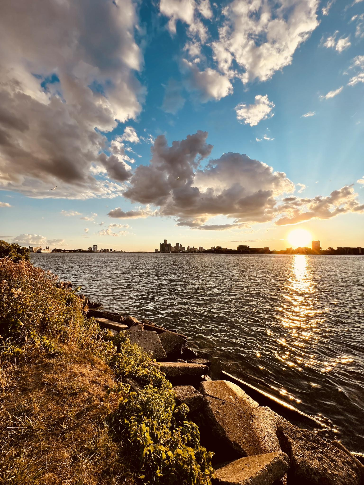 Golden sunset over water, rocky shore, and a distant city skyline.