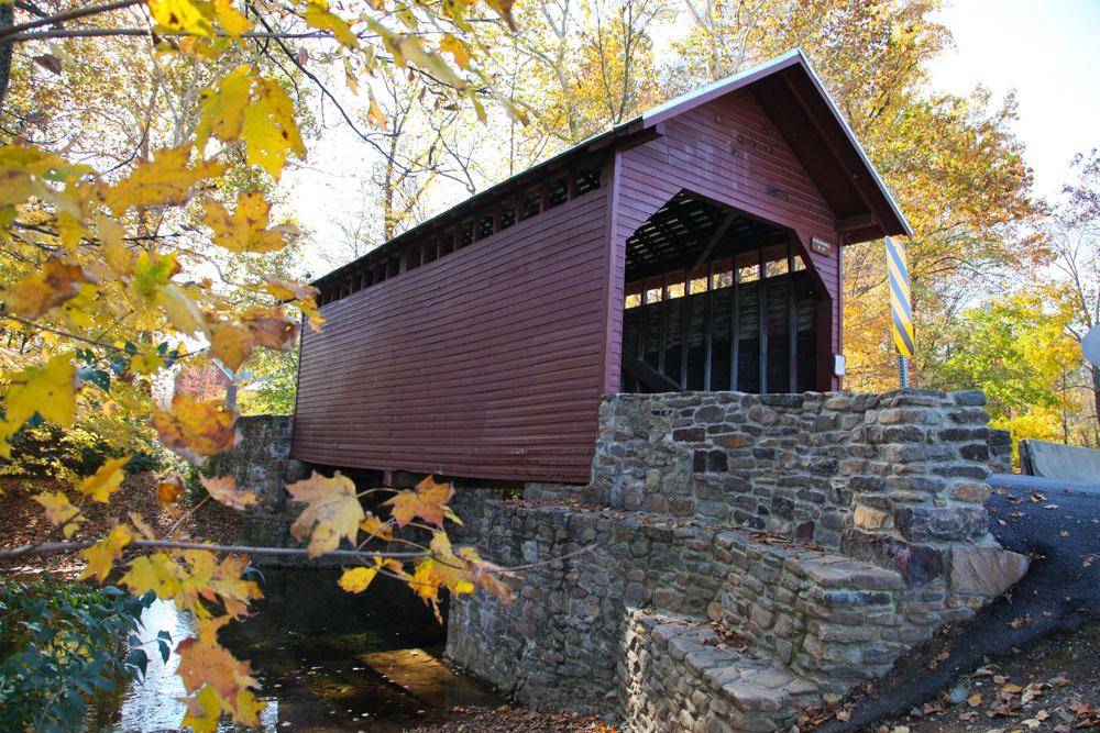 Red covered bridge over a stream, with bright yellow autumn leaves on a sunny day.