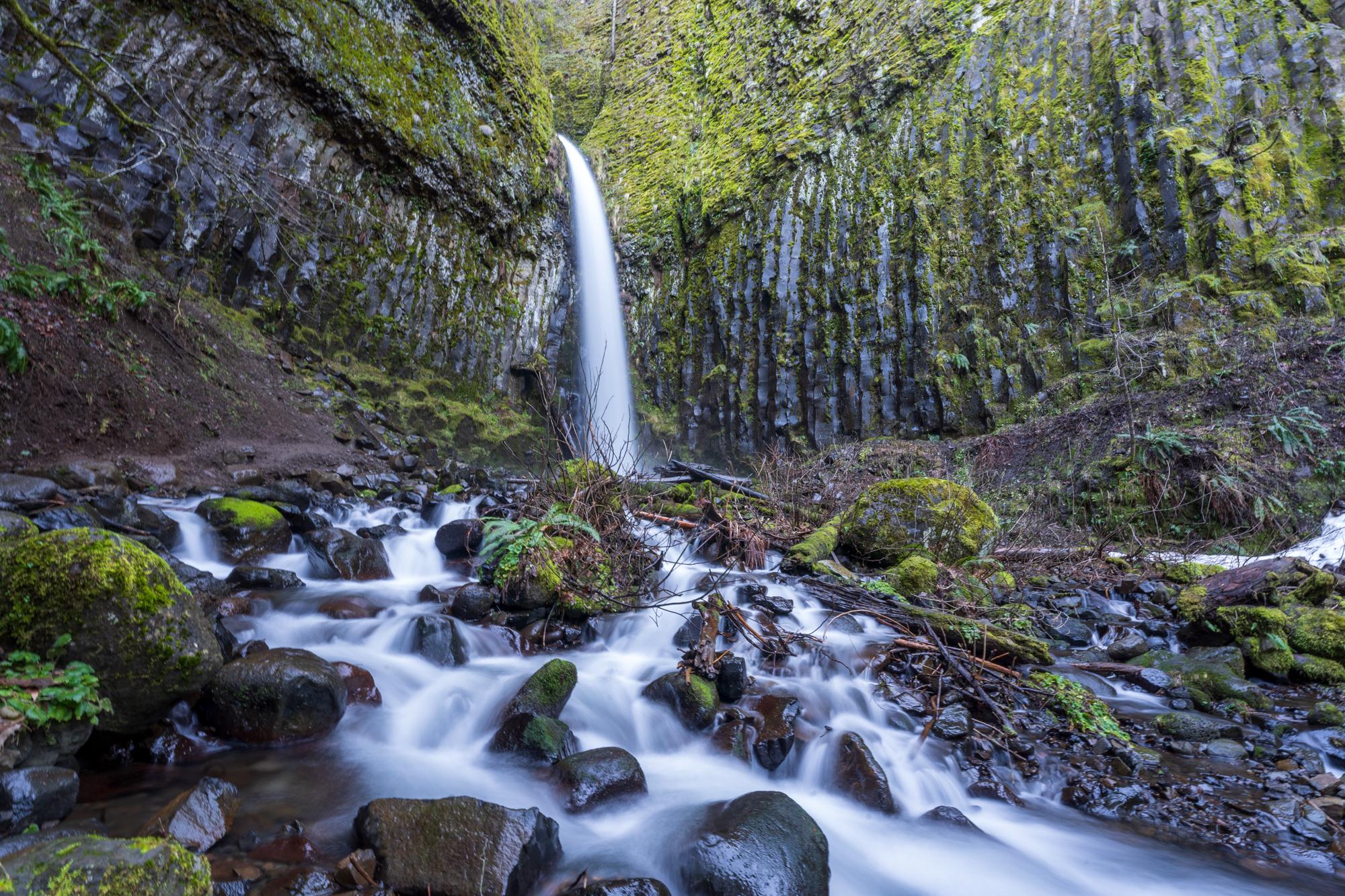 A powerful waterfall cascades over mossy columnar rocks into a rocky stream.