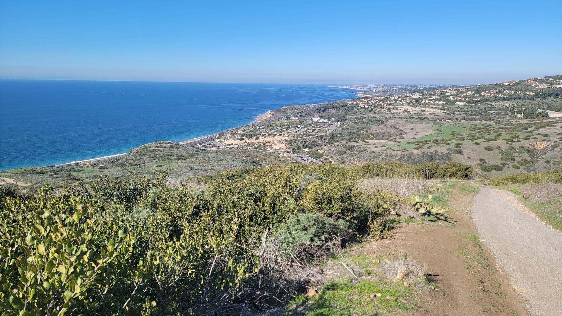 A wide view of the ocean coastline with green hills and a dirt path.