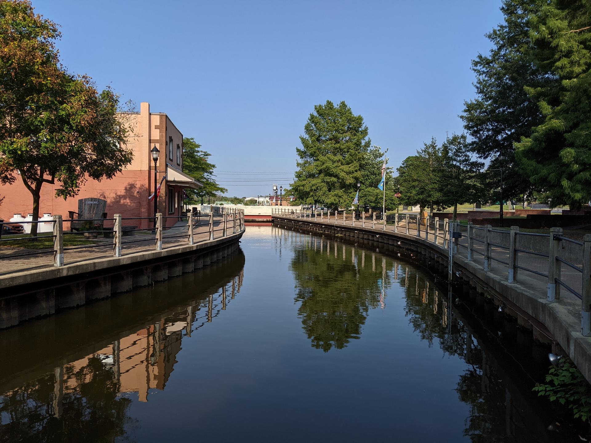 A sunny canal with paved walkways, buildings, and green trees along its banks.