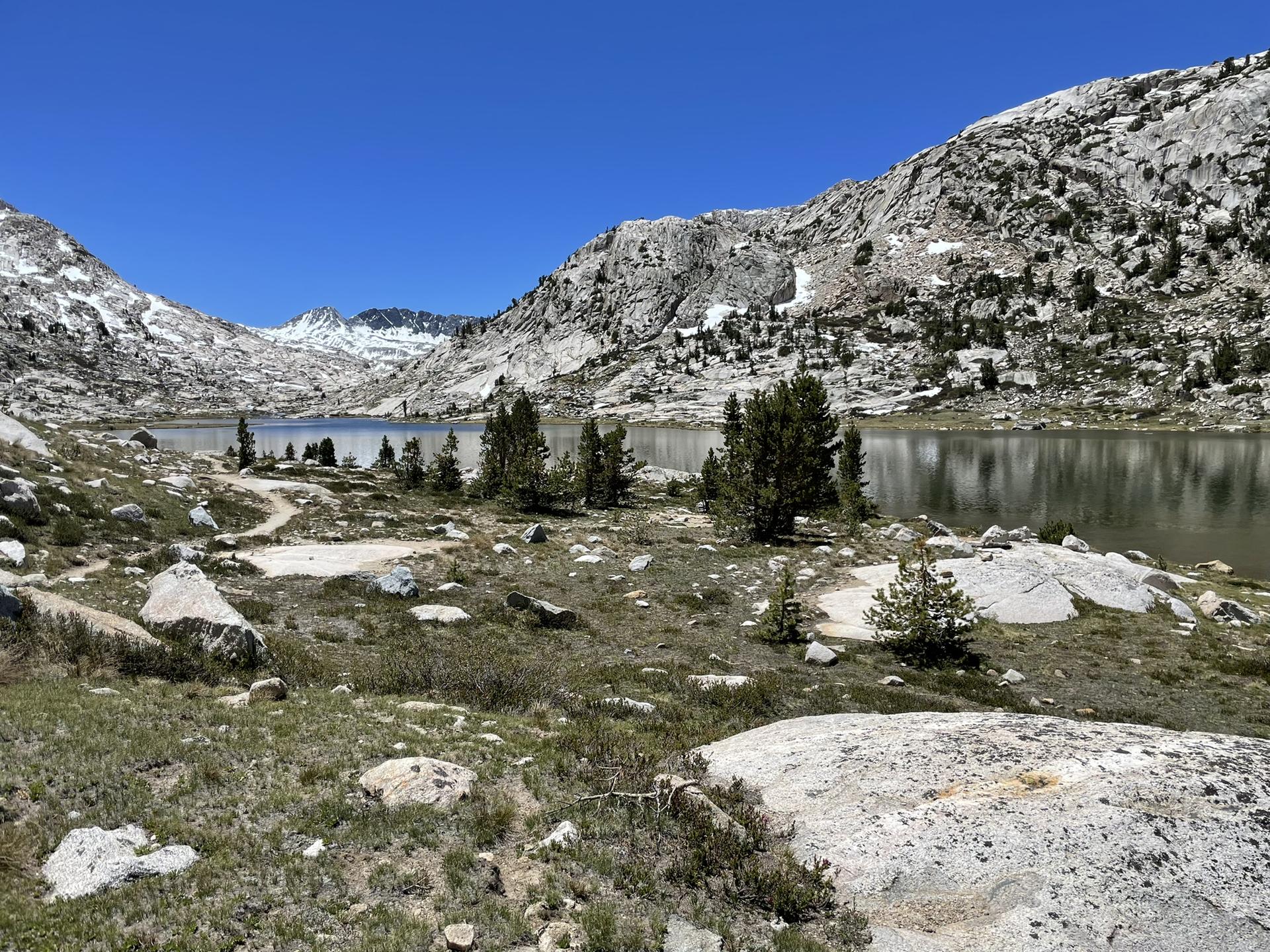 A mountain lake with rocky shores, sparse trees, and distant snow-dusted peaks.