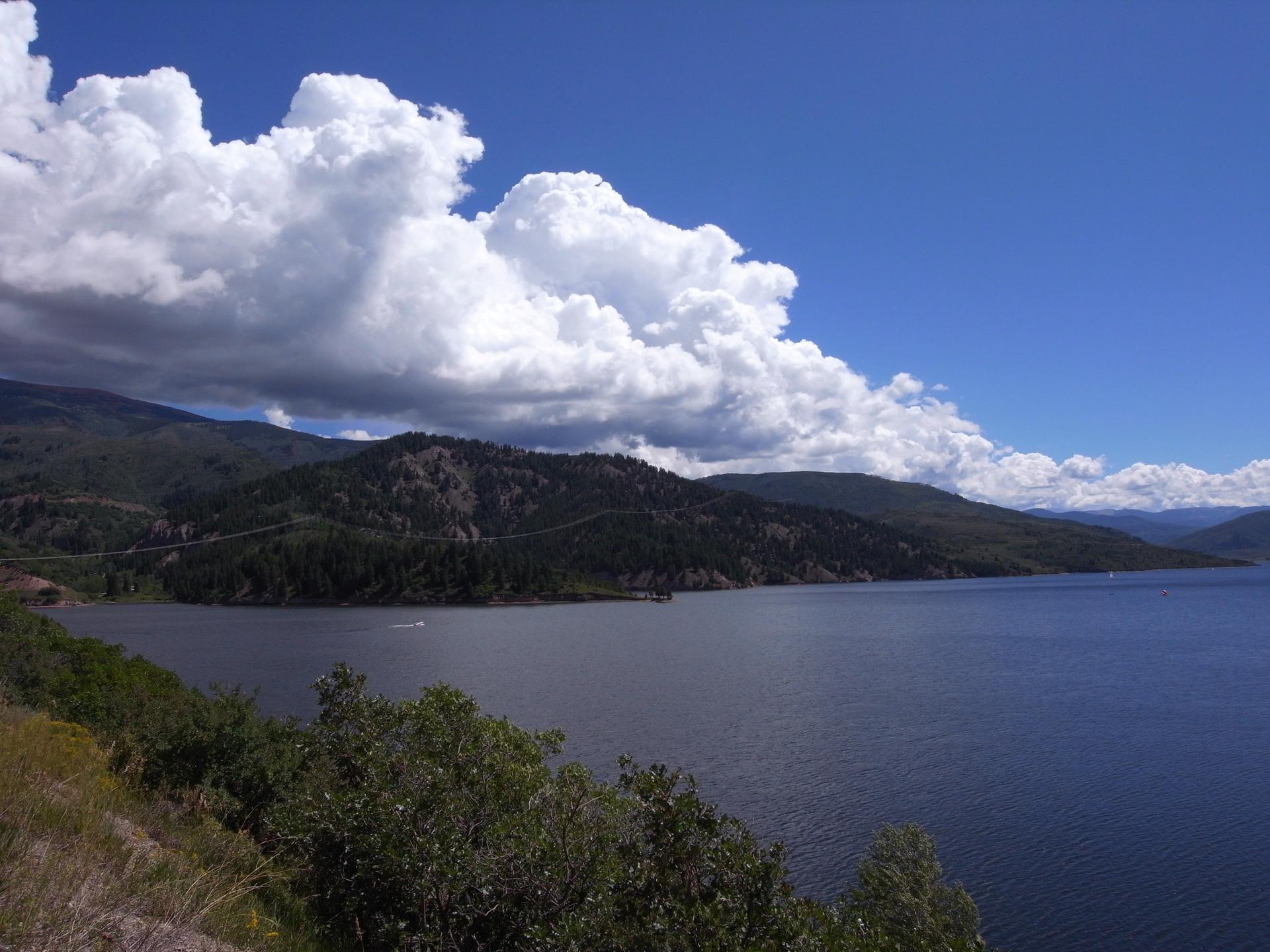 Expansive lake surrounded by forested mountains under a bright, cloudy sky with a boat.