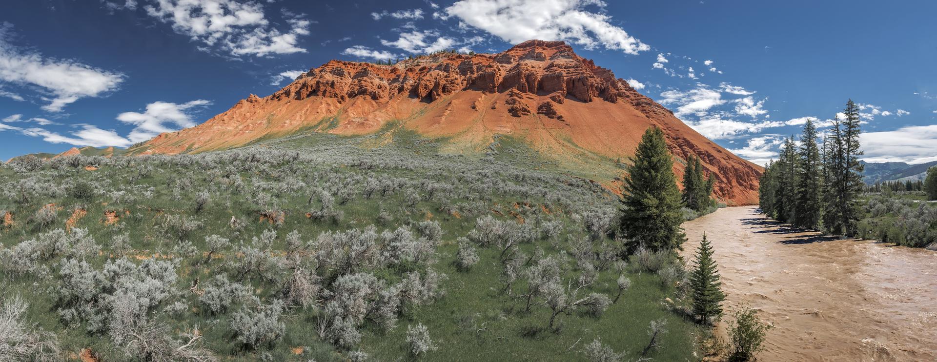 Panoramic view of a large red mountain, river, and green brush under a blue sky.