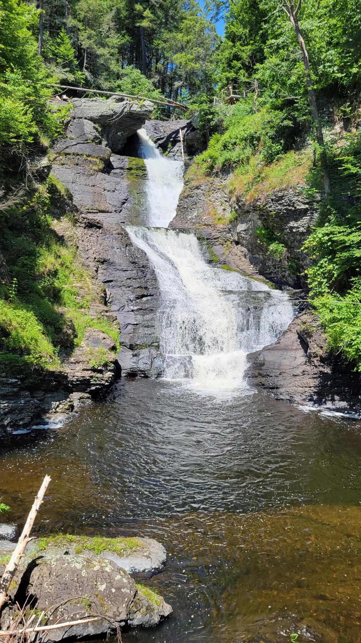 Multi-tiered waterfall cascading into a dark pool, surrounded by green trees.