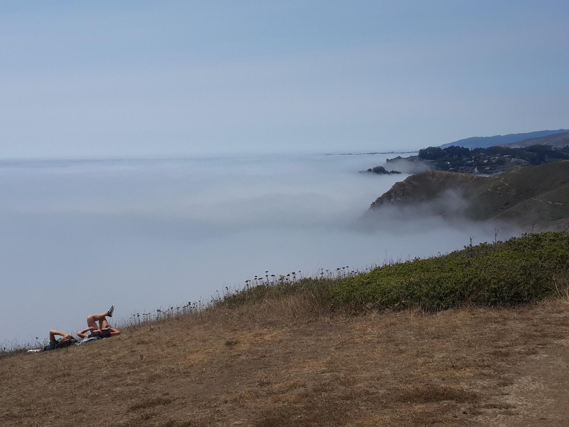 Two people relaxing on a grassy hill overlooking a foggy ocean.