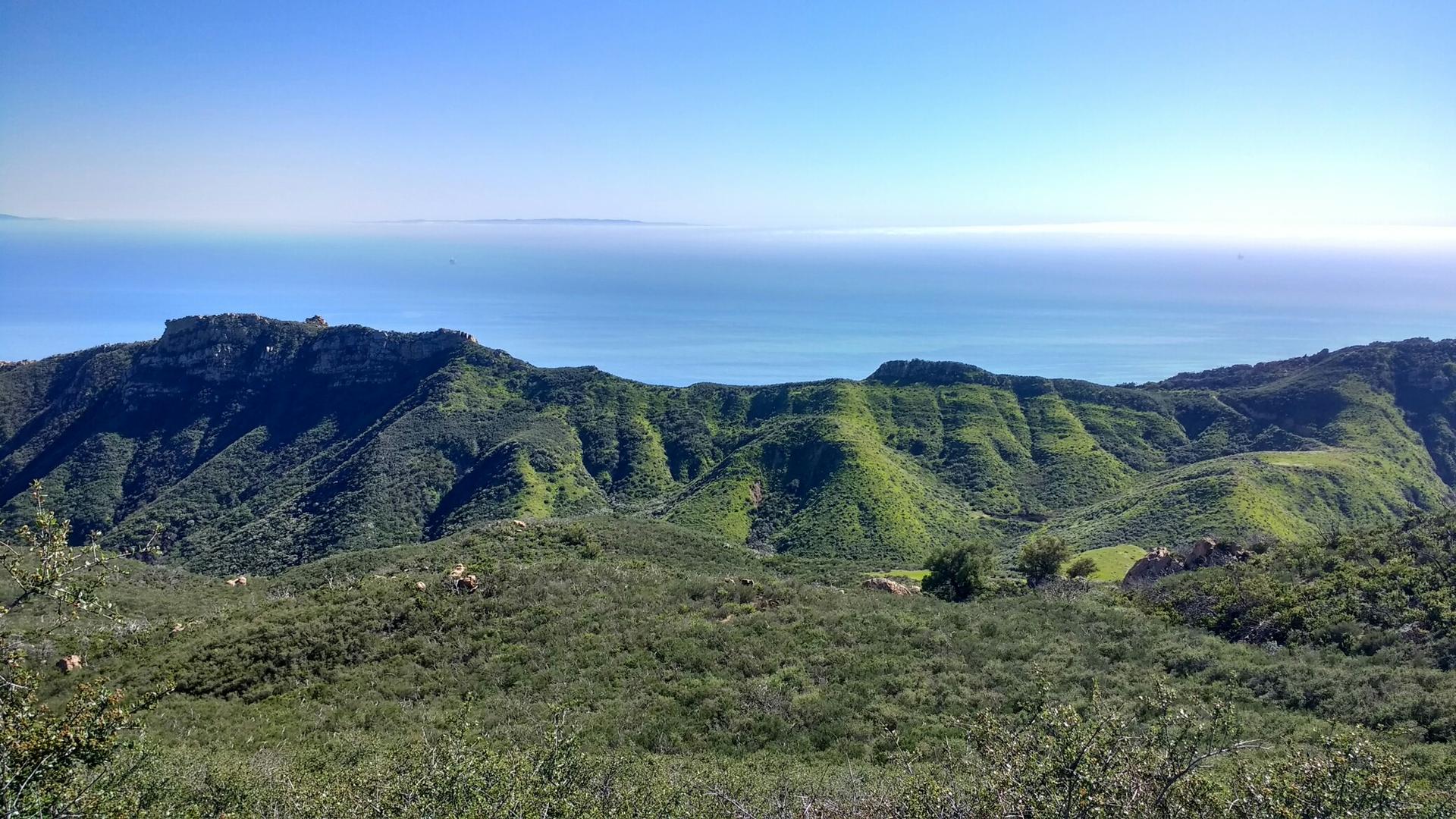 Expansive view of green mountains meeting the ocean under a bright blue sky.