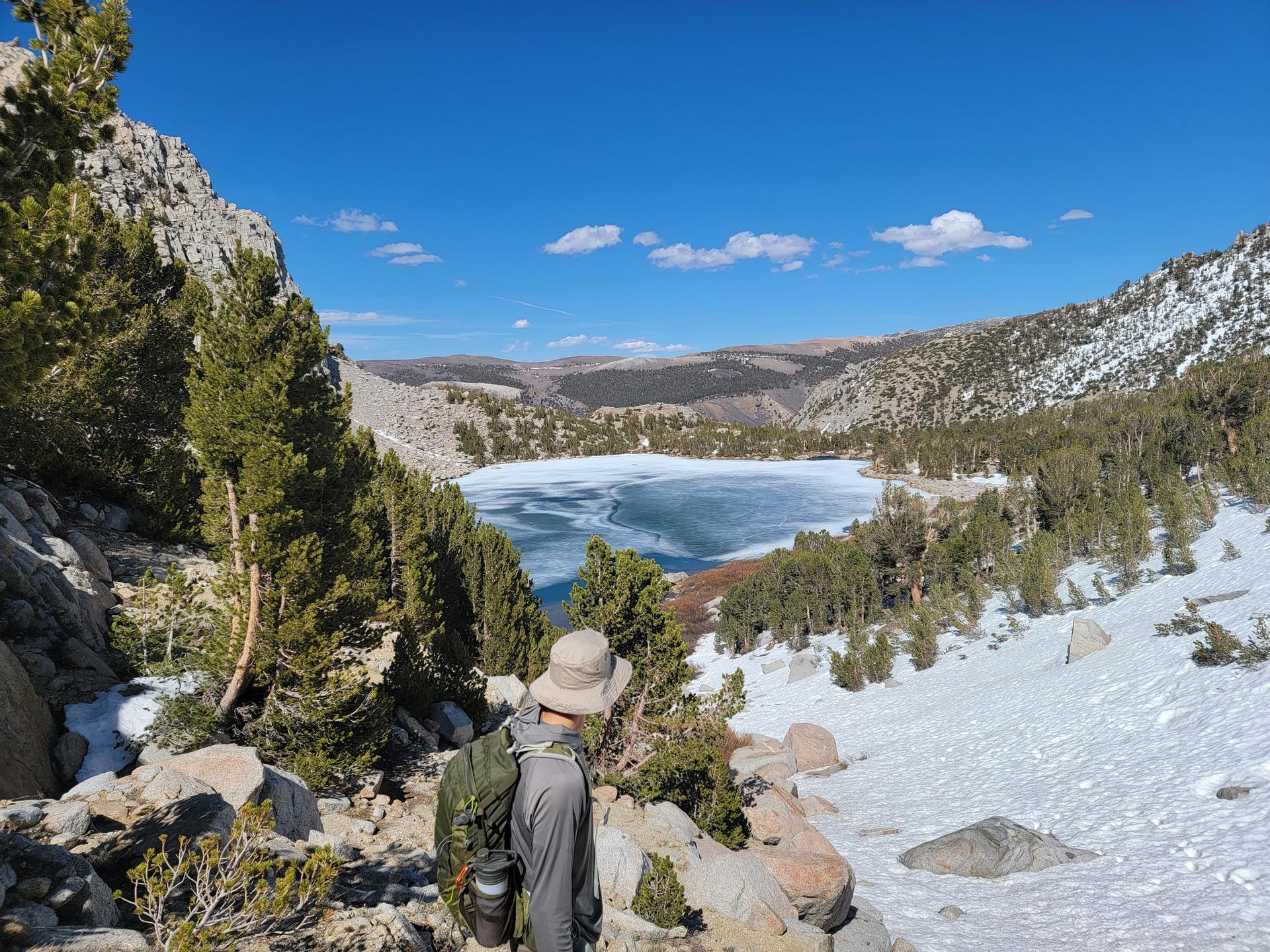 Hiker views a partially frozen lake surrounded by snowy mountains under a clear sky.