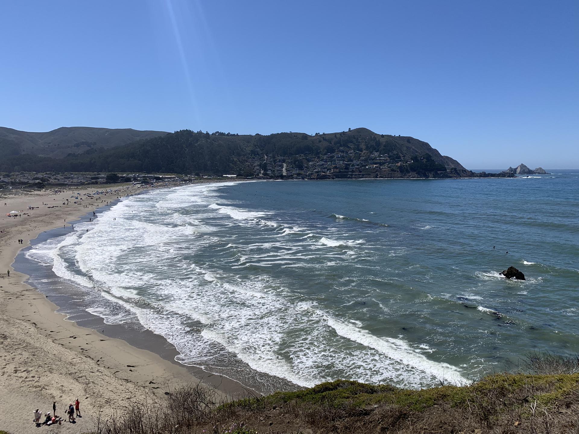 Wide sunny beach with many people, crashing waves, and distant hills.