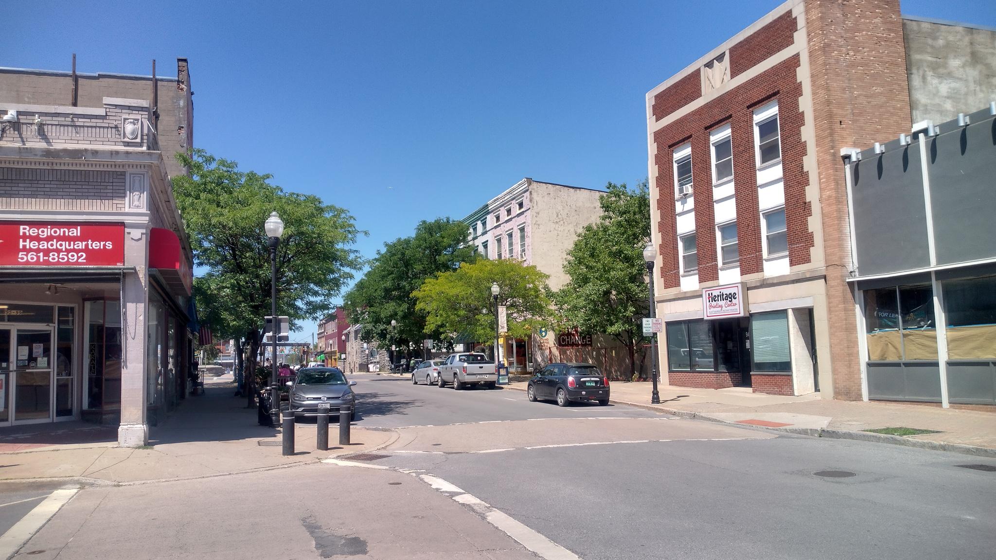 Sunny urban street with brick buildings, parked cars, and green trees.