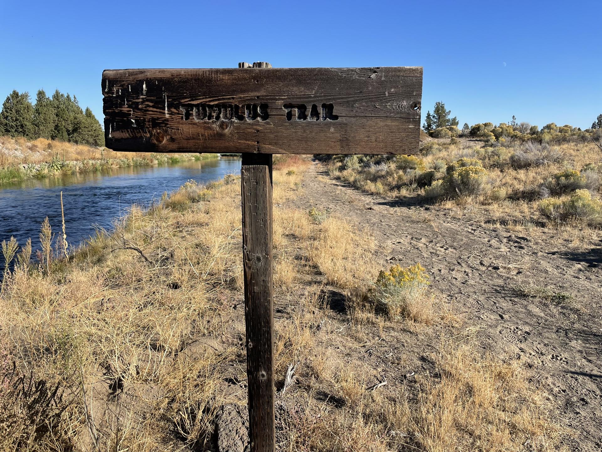 A rustic wooden trail sign stands beside a river and dry path.