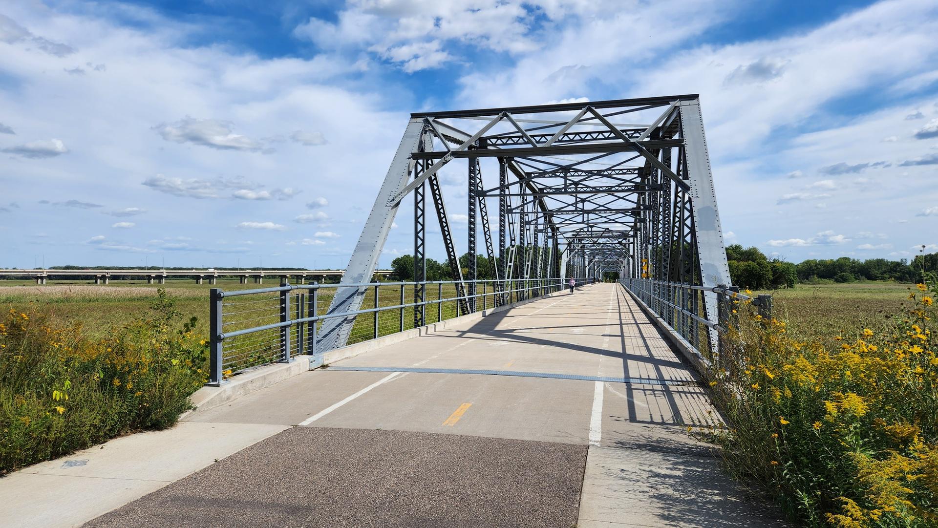 A large metal truss bridge spans over a paved path on a sunny day.