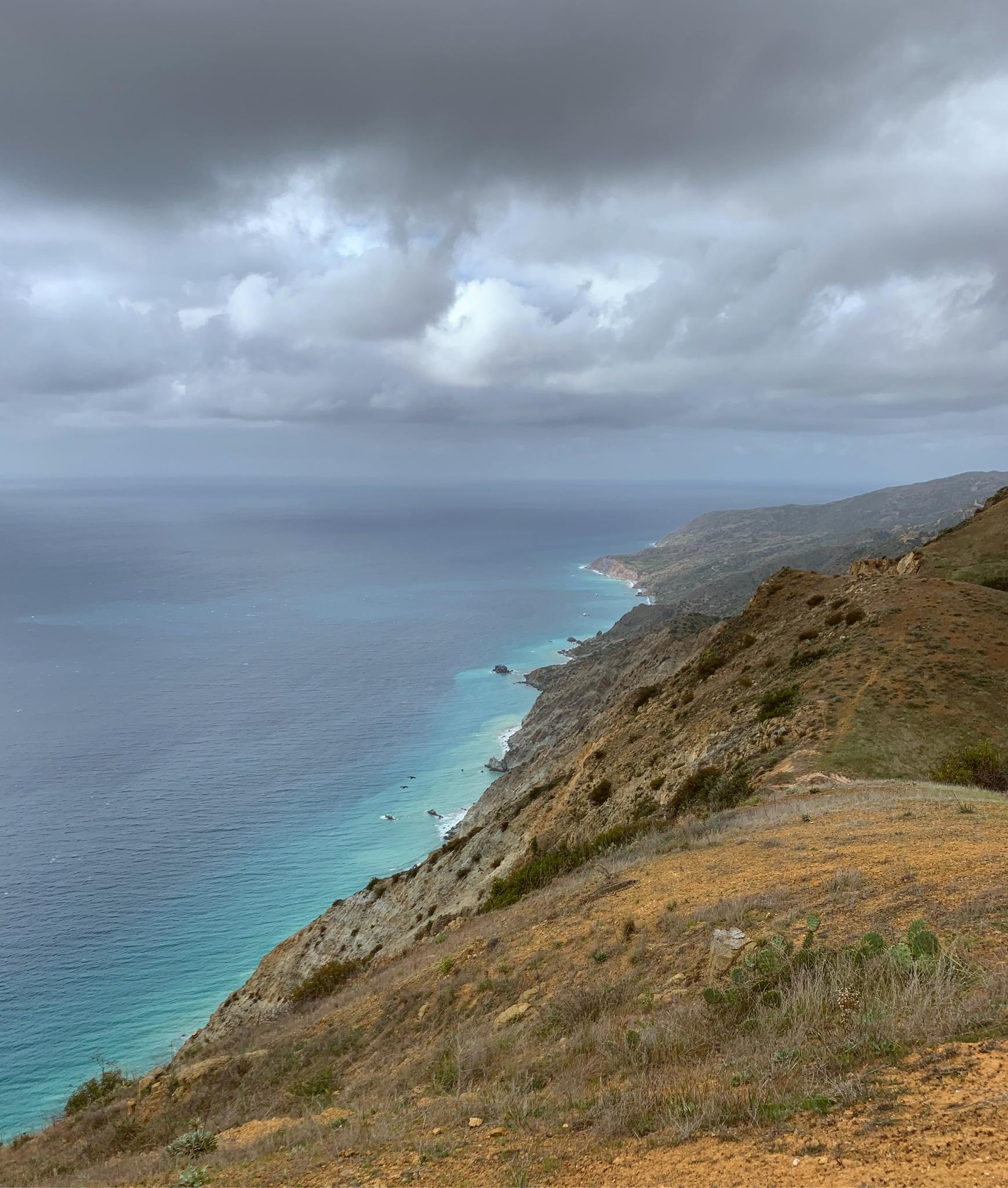 Steep, dry coastal cliffs overlook turquoise ocean under a cloudy sky.