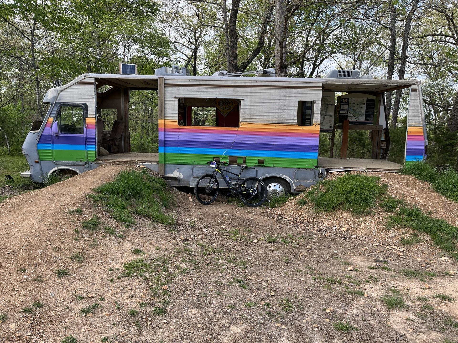 A rainbow-painted RV converted into an open structure, with a bicycle nearby.