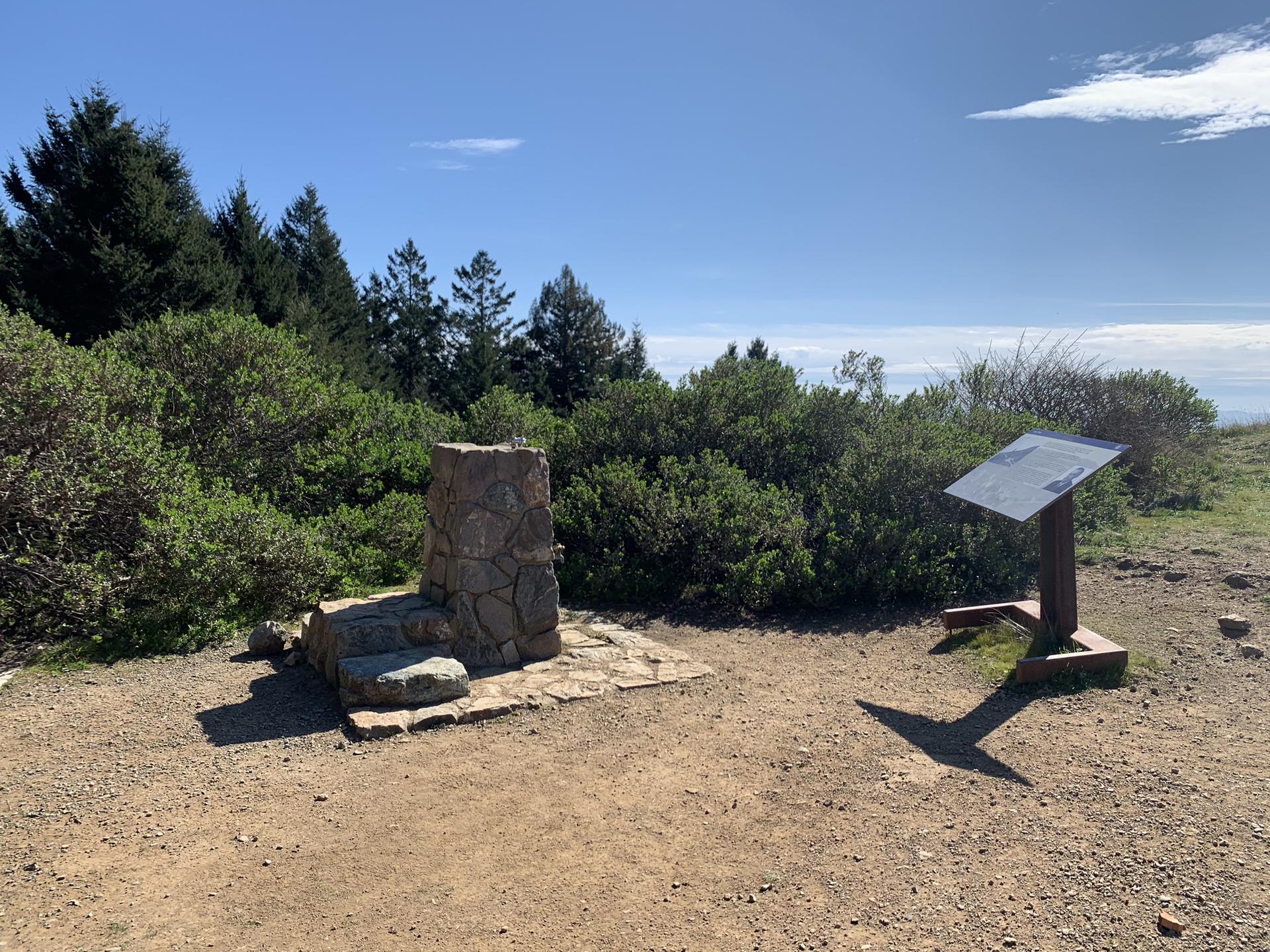Stone monument and interpretive sign on a sunny dirt clearing.