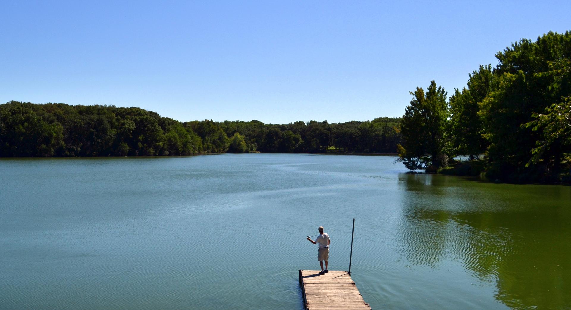 Man on a wooden dock overlooking a wide lake surrounded by green trees.