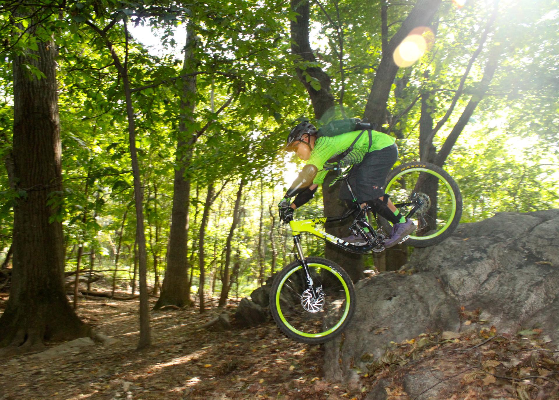 Mountain biker in green rides over a large rock in a sunny forest.