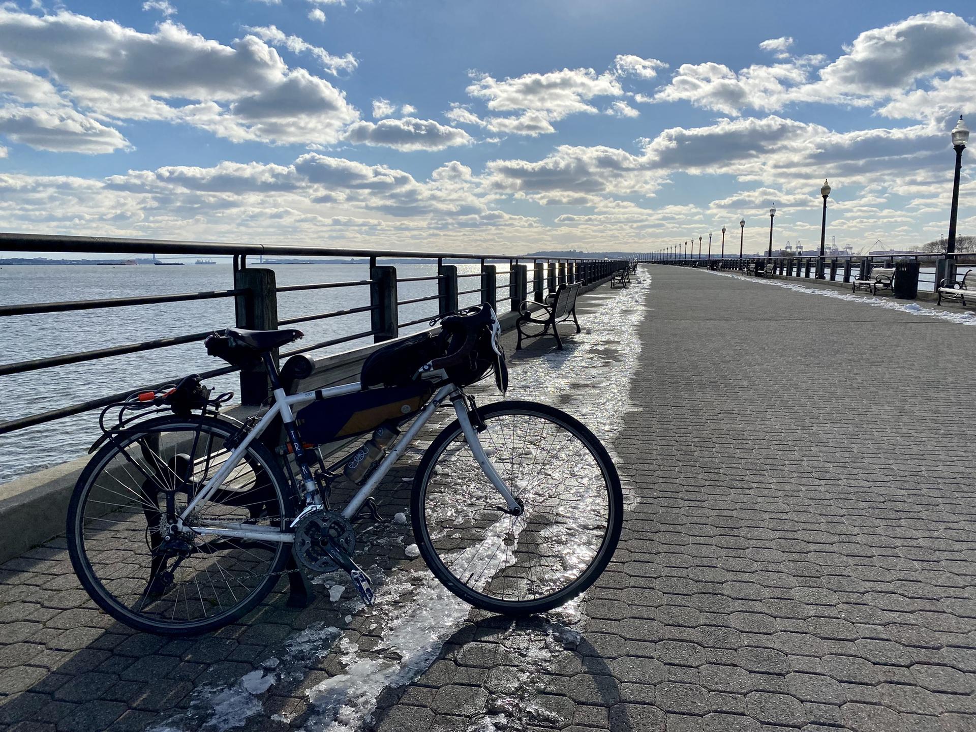 A bicycle parked on a sunny waterfront boardwalk with patches of snow.