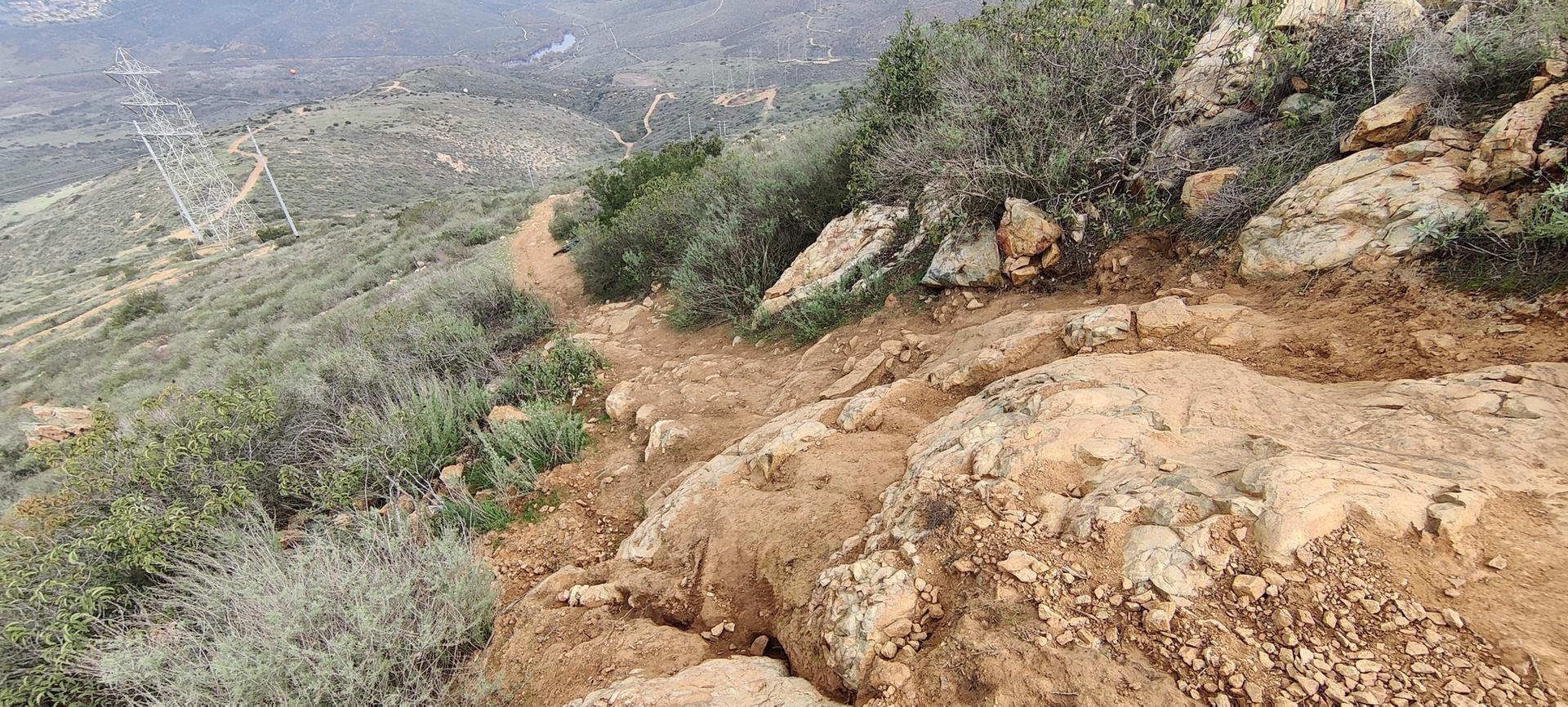 A steep, rocky dirt trail winds down a brush-covered hillside.