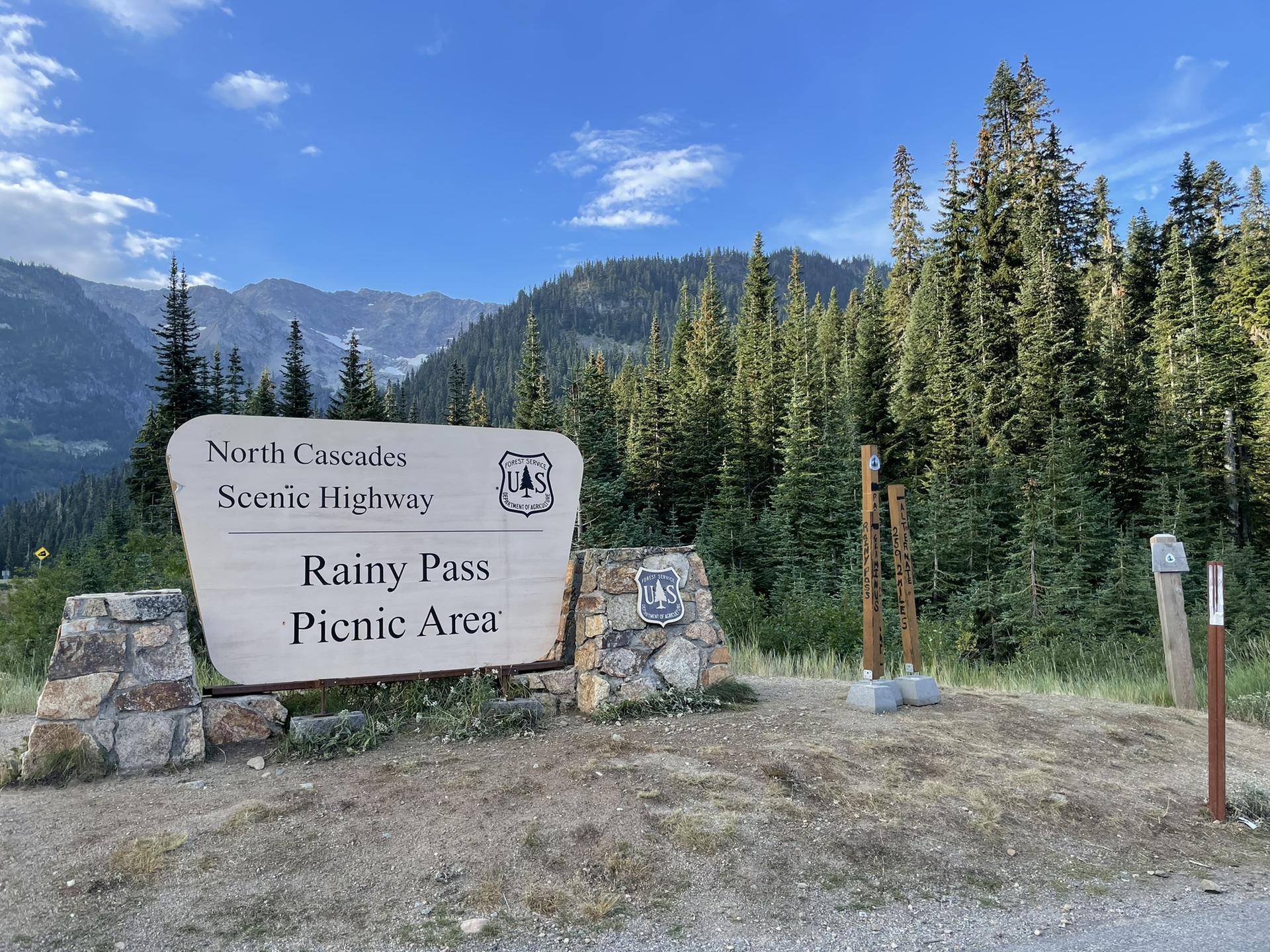 A large wooden sign on a stone base in front of forested mountains.
