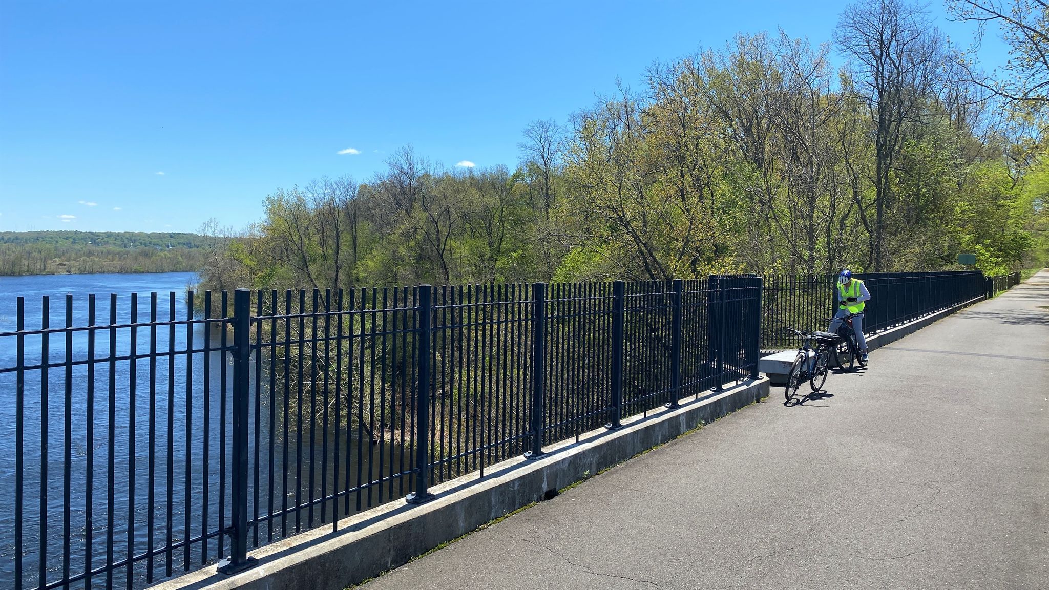 Cyclist stands by a black fence overlooking a wide river on a sunny day. - Farmington River Bridge – East Coast Greenway loop from Town Open Space (Sanford Drive)