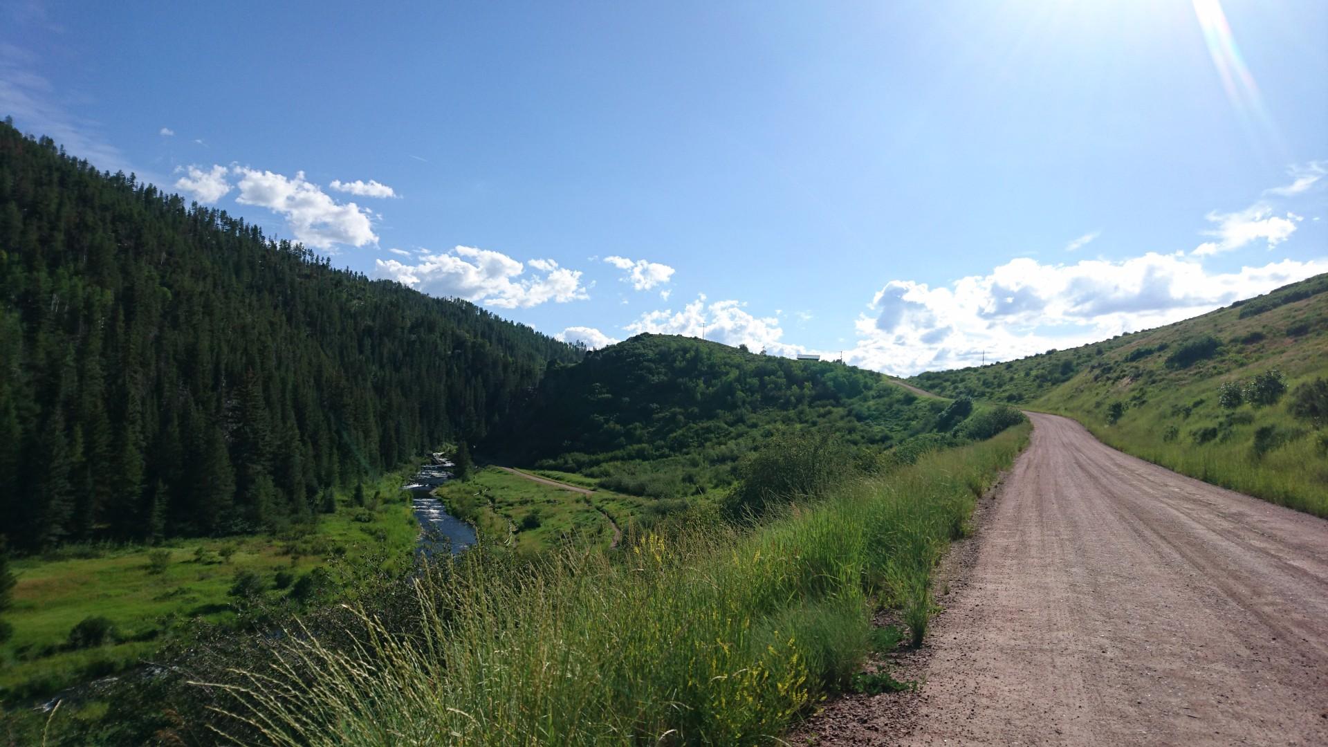 A dirt road winds alongside a green valley with a stream under a bright sky.