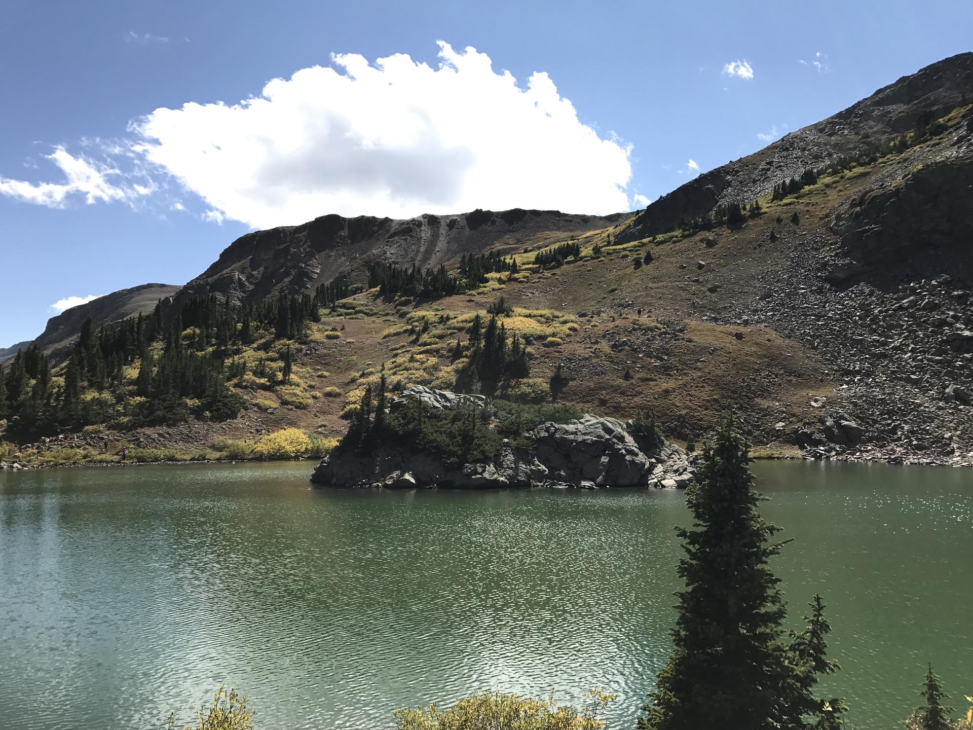 Serene mountain lake with a rocky island and steep, forested slopes under clouds.