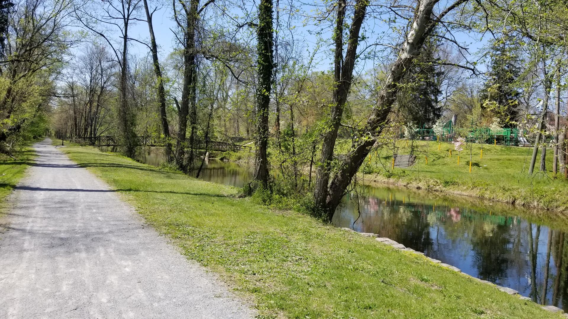 A gravel path runs beside a narrow canal with green trees and grass.