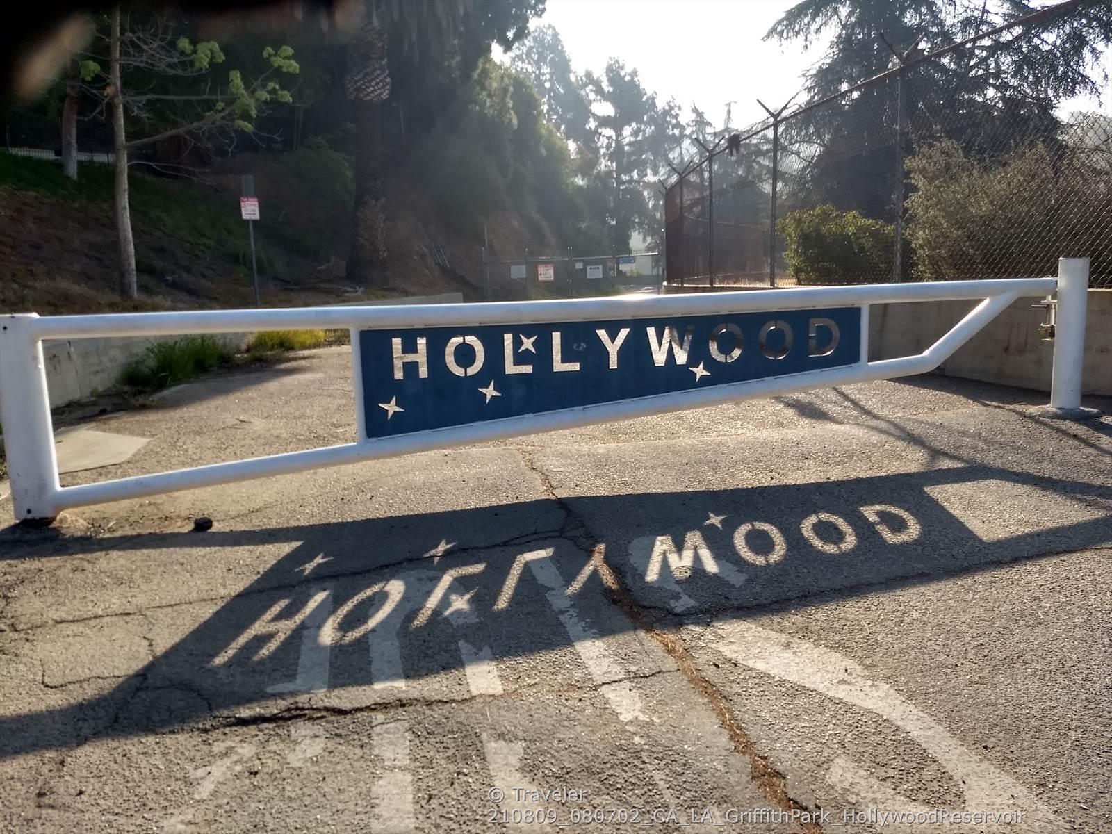 A white gate with a blue sign casts a long shadow on a paved road.