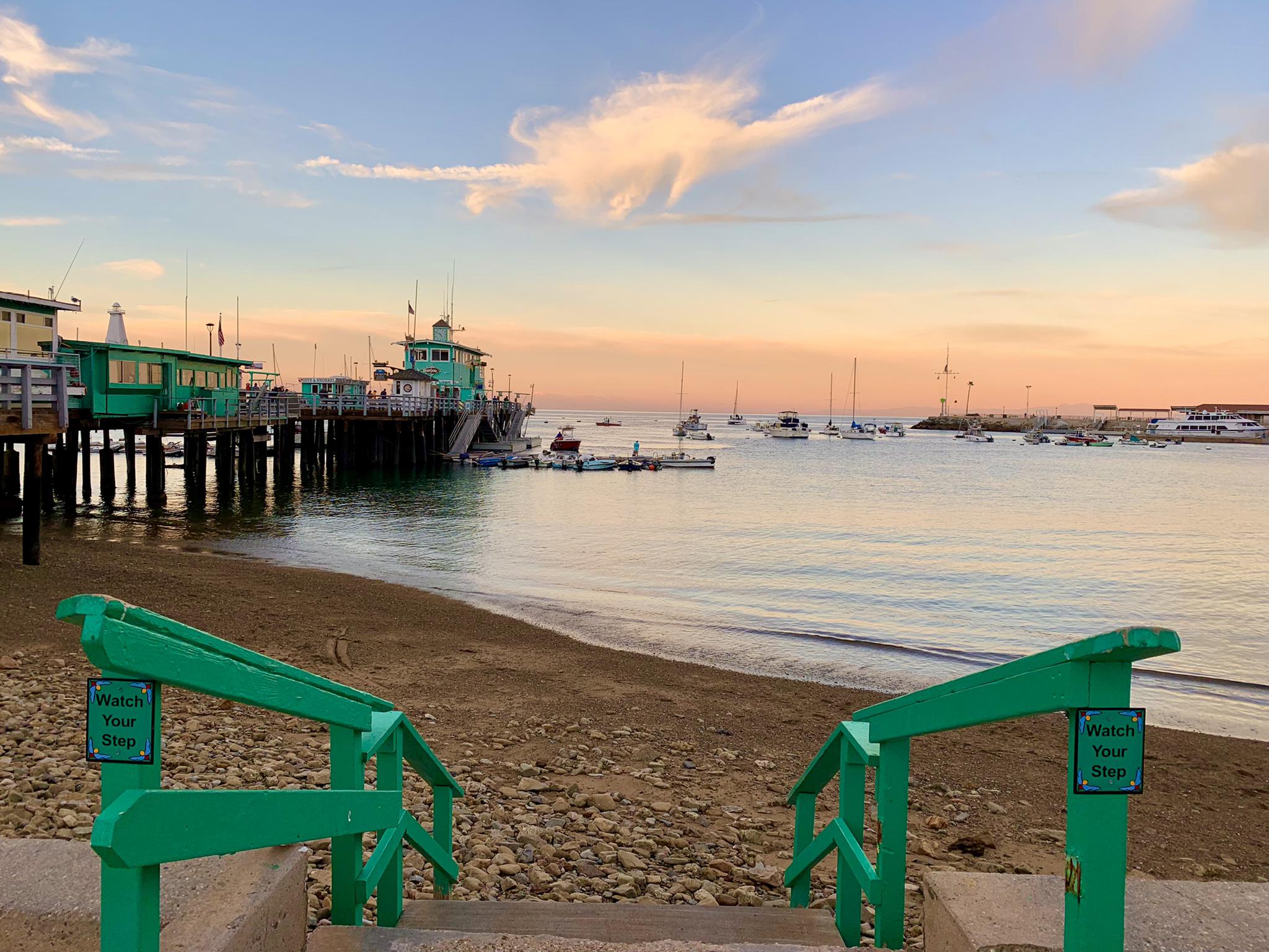 Green railings lead to beach, pier, and boats at sunset.