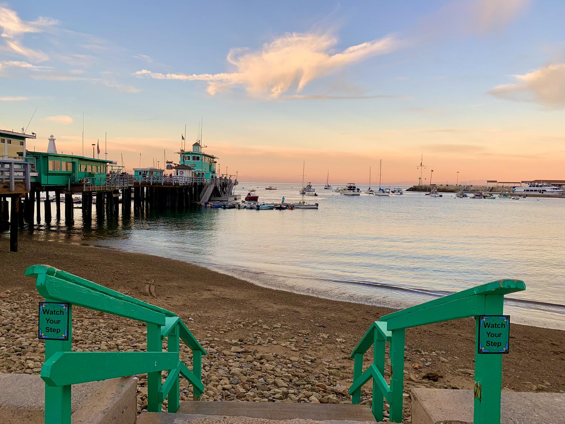 Green railings lead to beach, pier, and boats at sunset.