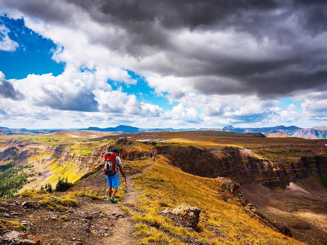 Hiker with backpack walks along a vast, golden mountain ridge under cloudy skies.
