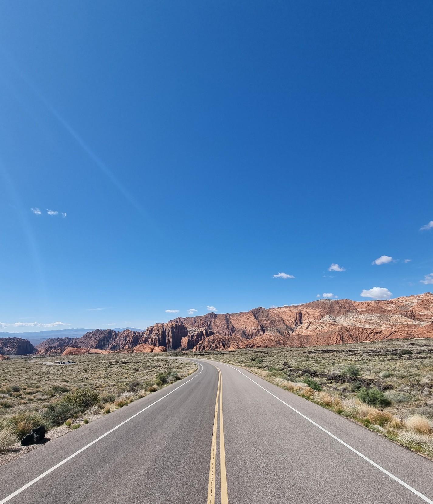 A winding desert road leads towards red rock formations under a bright blue sky.