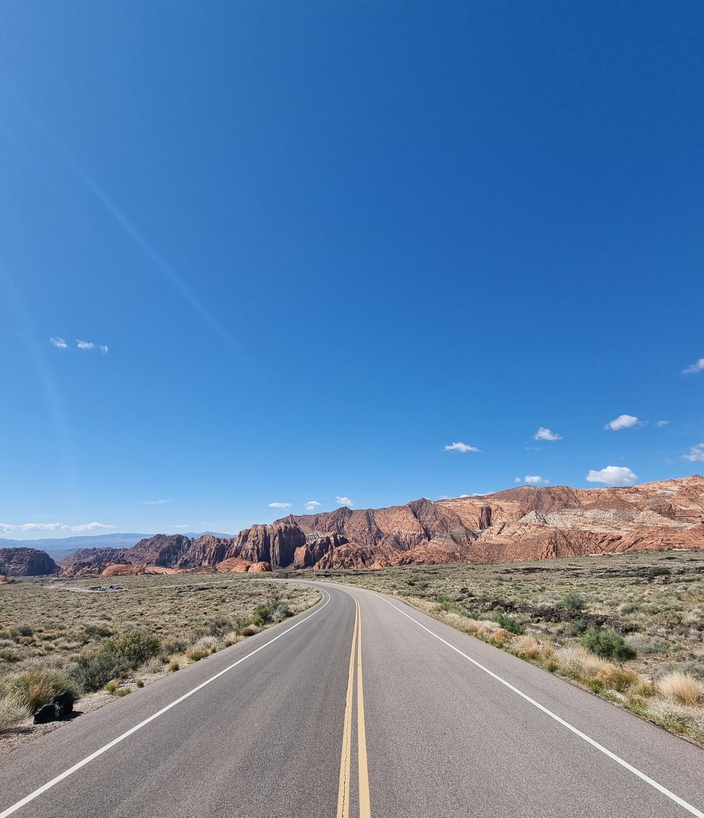 A winding desert road leads towards red rock formations under a bright blue sky. - Snow Canyon & Gunlock Reservoir State Parks loop