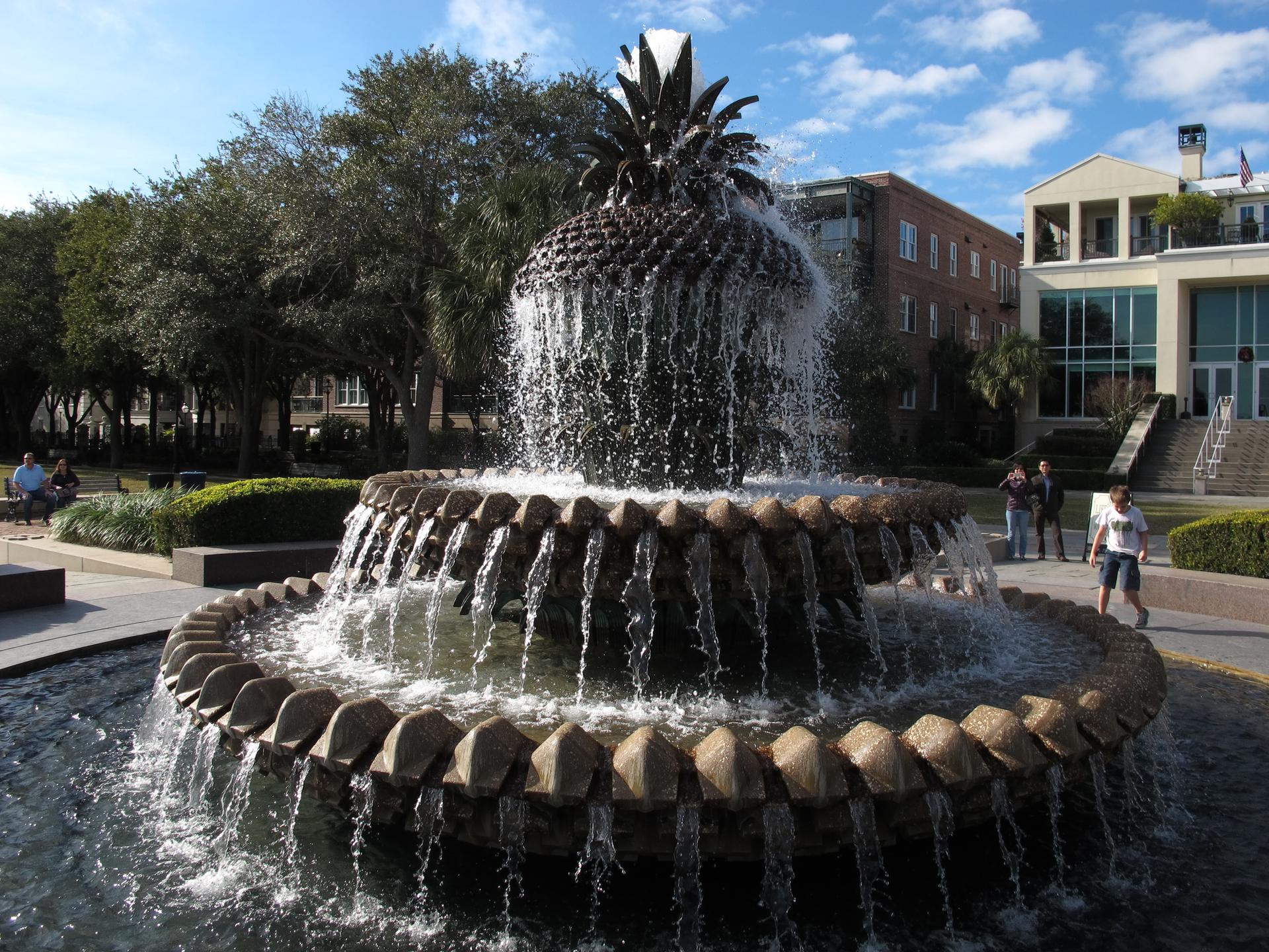 Large pineapple fountain spraying water in a sunny park with people.