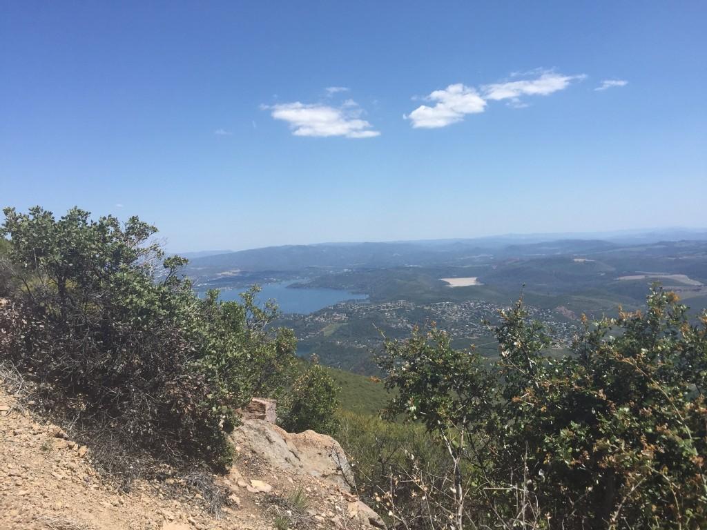 Sunny panoramic view of a large lake surrounded by green hills and distant mountains.