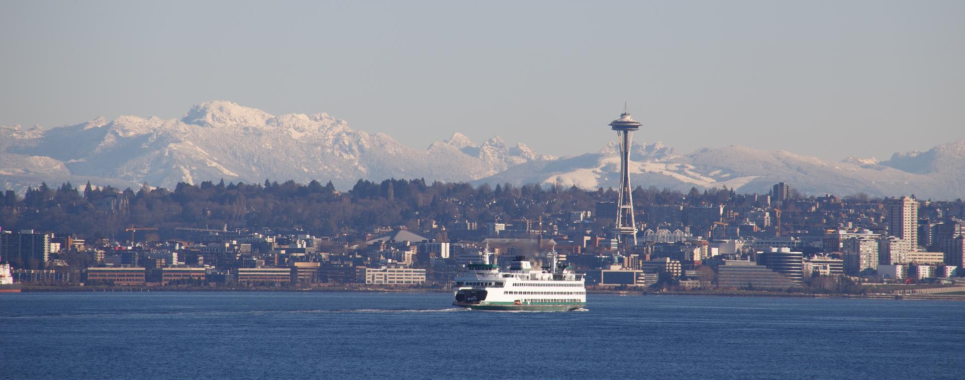 Ferry boat on blue water, city skyline, and snow-capped mountains under clear sky.