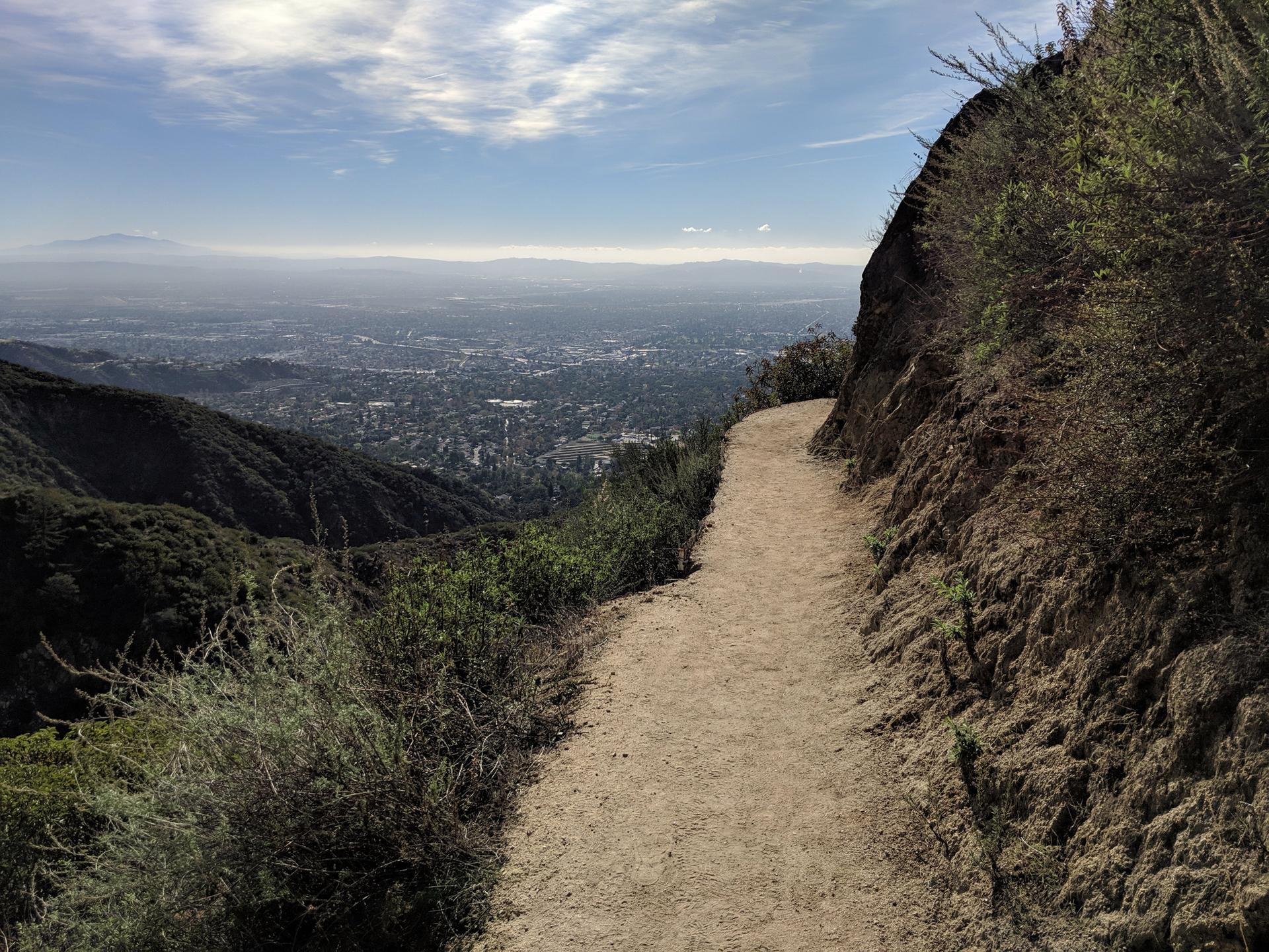 Dirt path along a steep hillside overlooking a city under hazy sky.