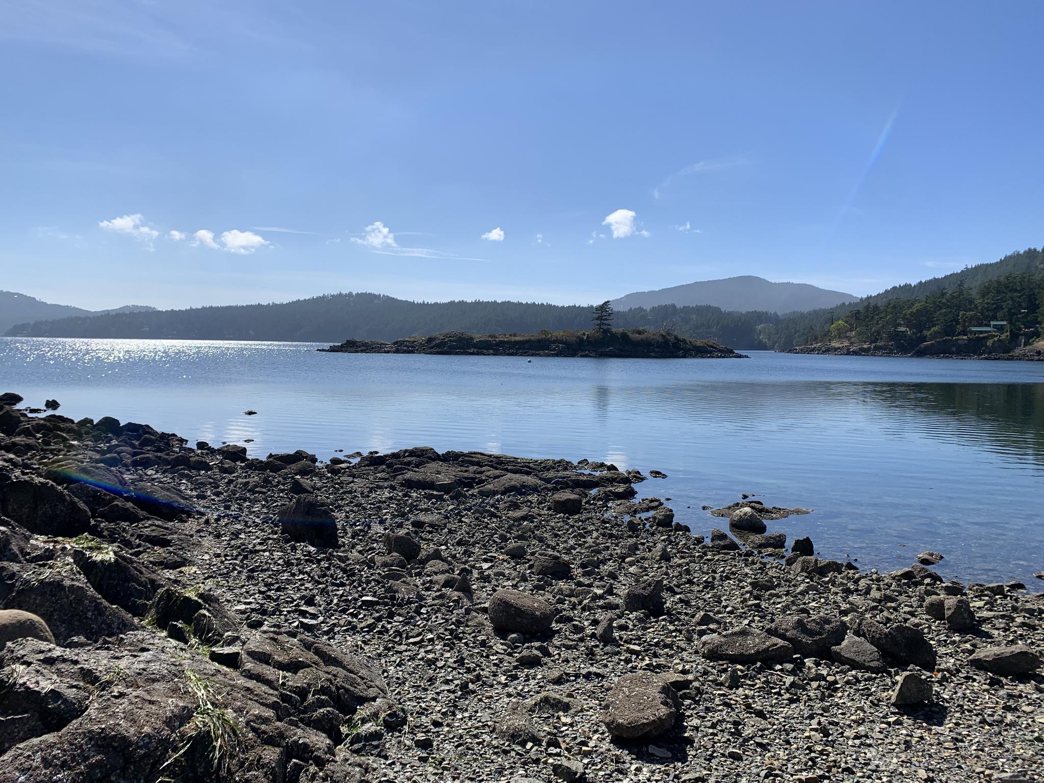 Rocky shoreline with calm blue water, distant forested islands under a bright sky.